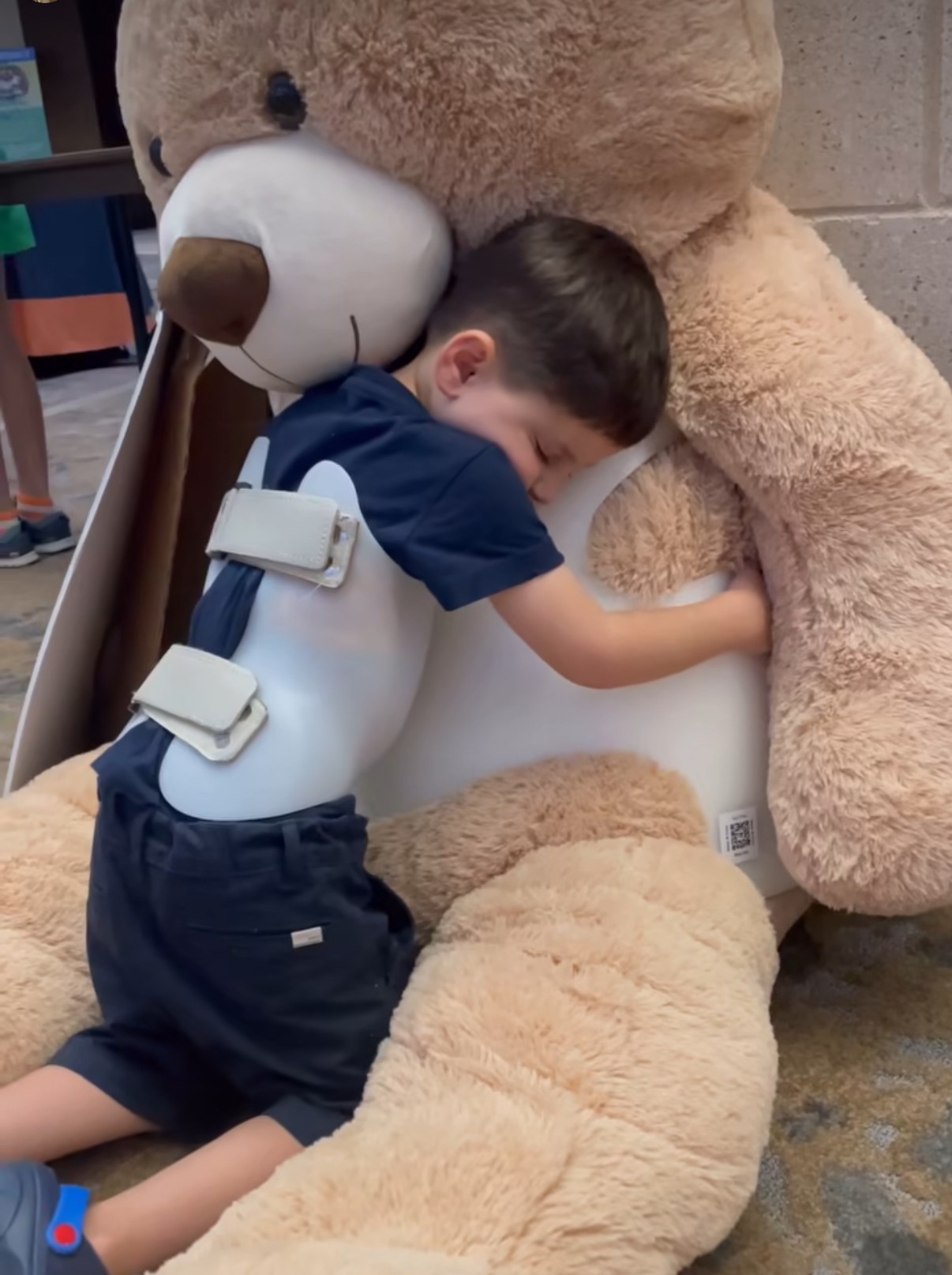 Child hugging a large teddy bear, with arms around its neck, in a room with a concrete wall.