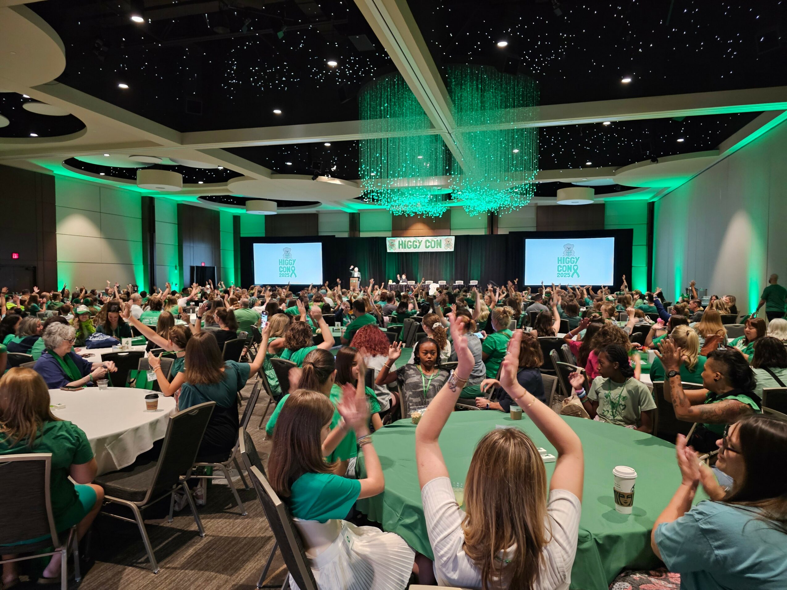 Large conference room filled with people seated at round tables, stage with screens, and green lighting, ceiling with star-like lights.