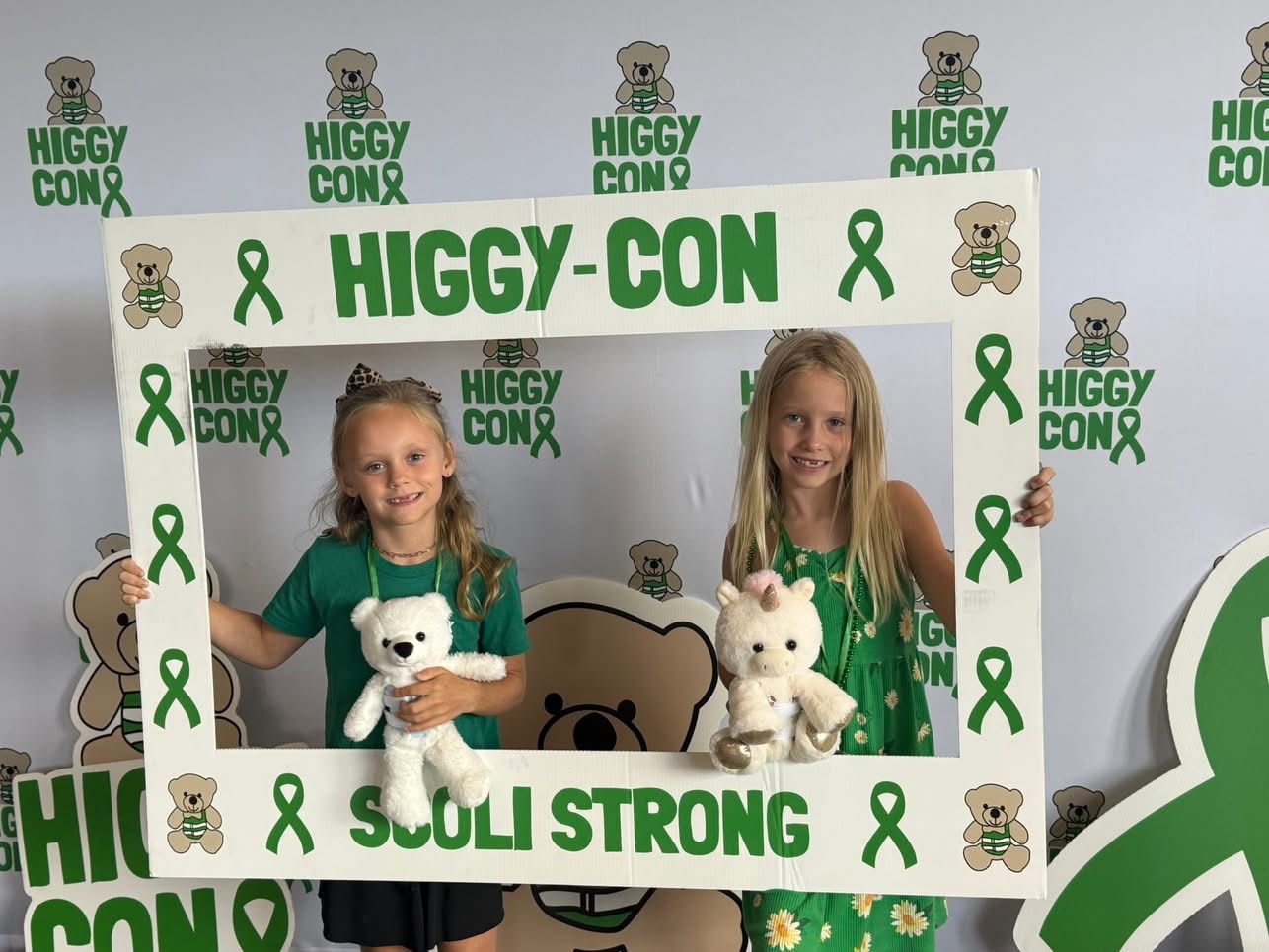 Two young girls in green shirts holding white teddy bears, standing behind a frame with green ribbons and text, at Higgy-Con.