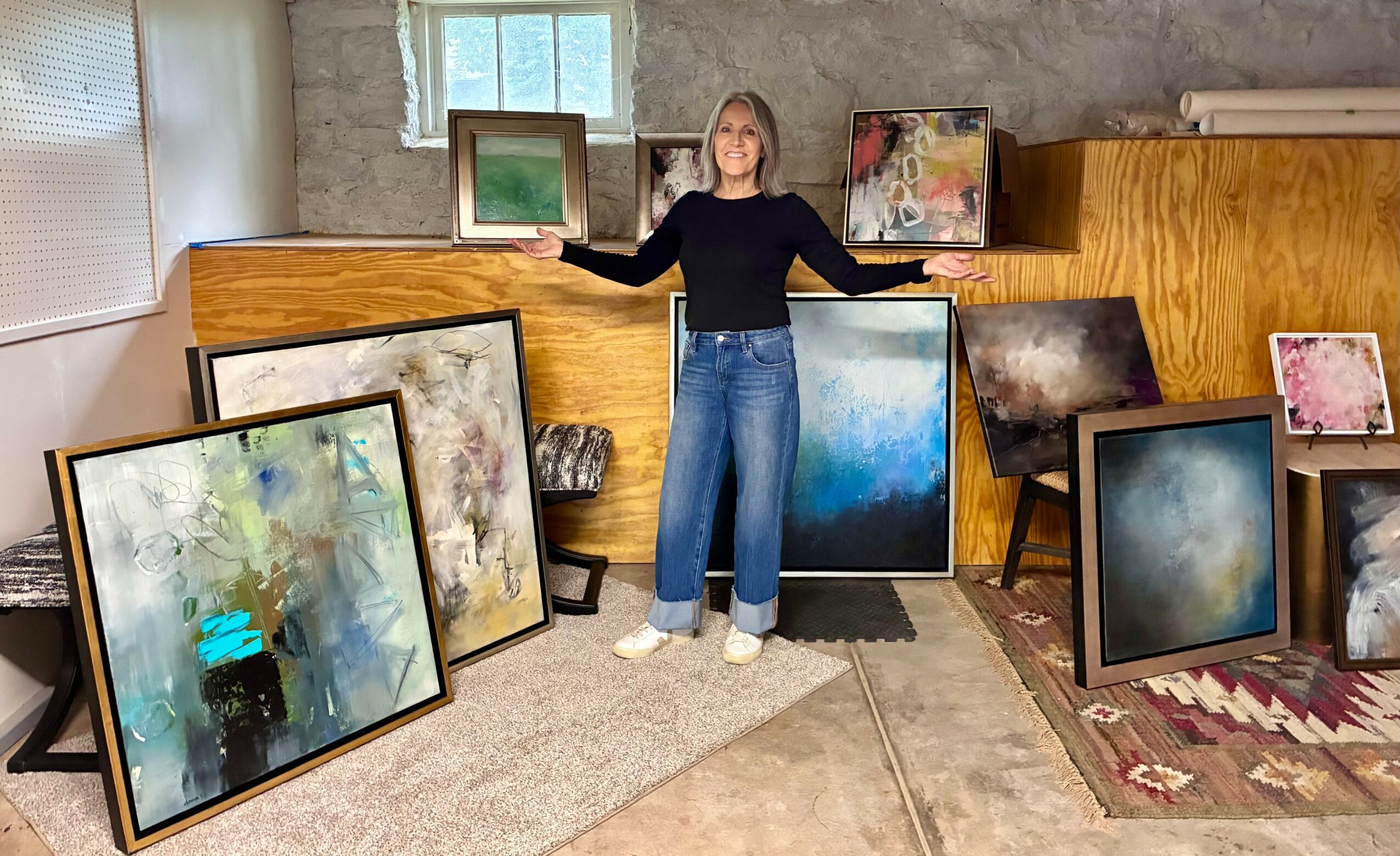 Person standing in art studio surrounded by framed abstract paintings on the floor and on a wooden shelf.