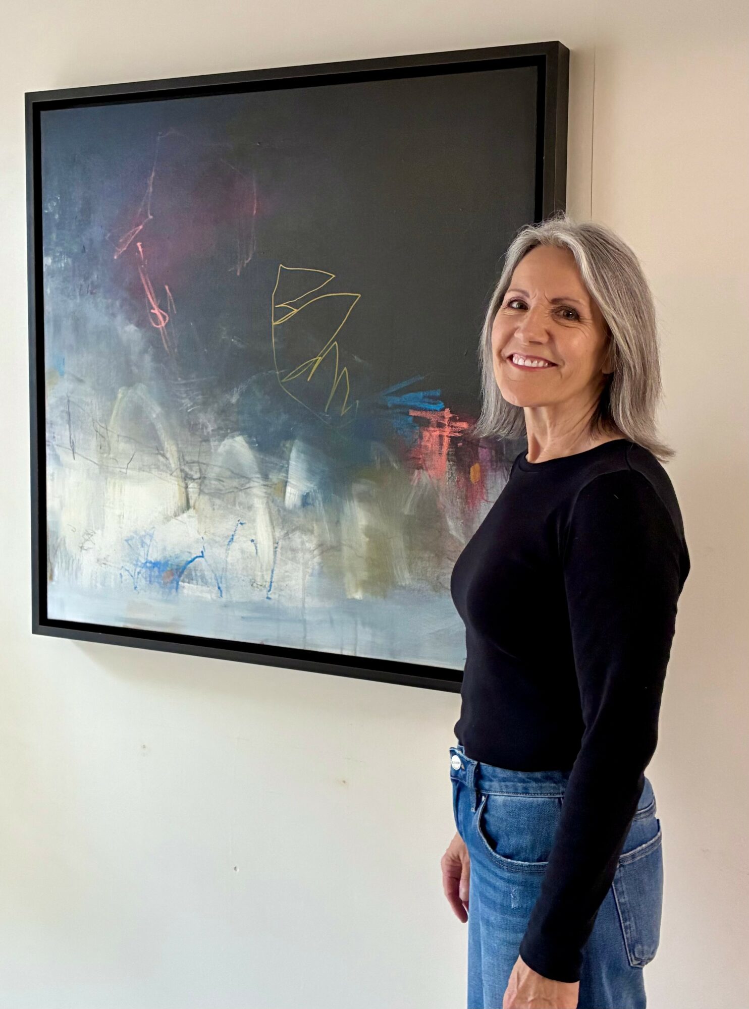 Smiling woman with gray hair wearing a black top and blue jeans standing next to an abstract painting on a white wall.
