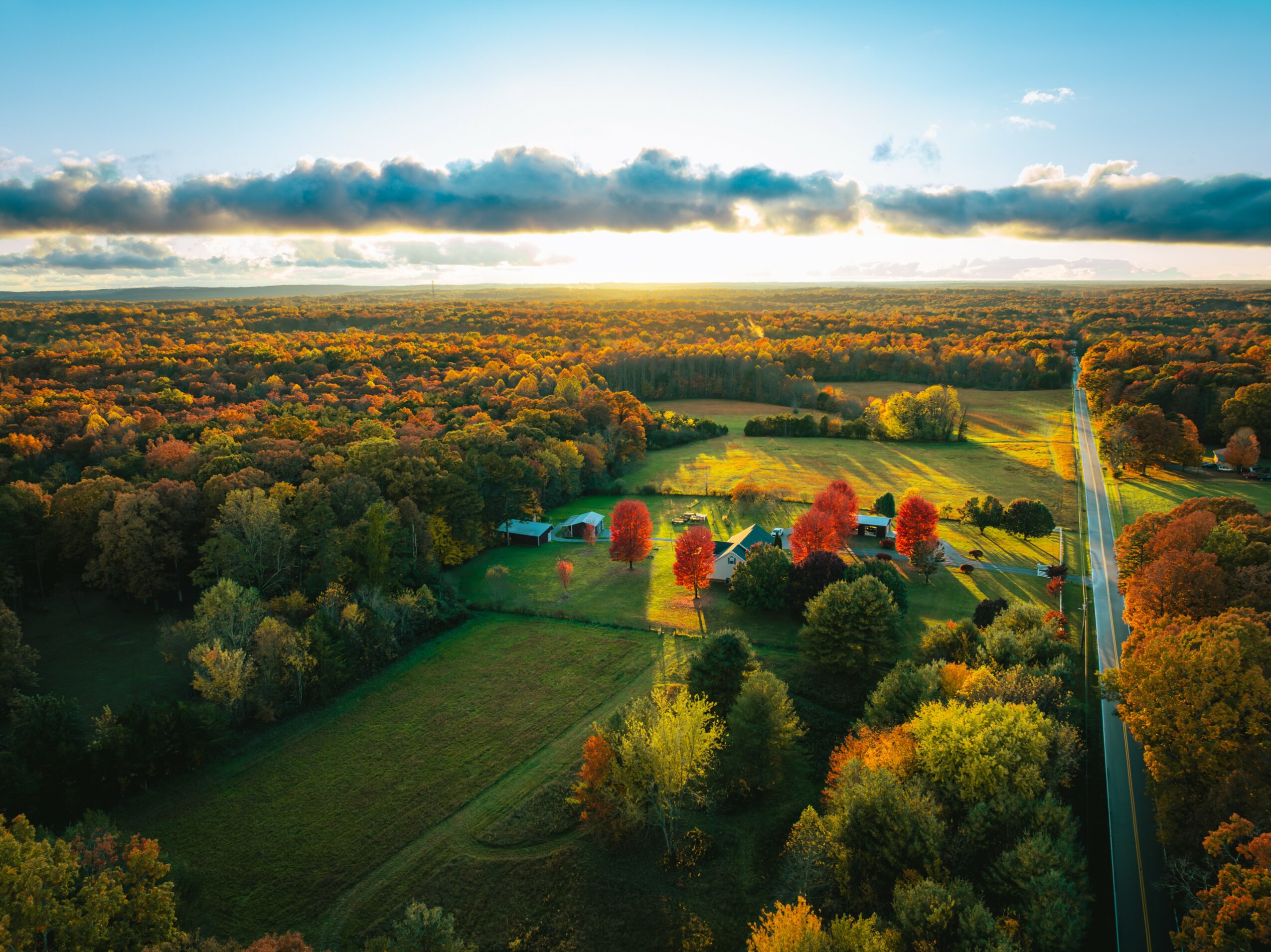 Aerial view of a landscape with colorful trees, open fields, a road, and a cloudy sky.