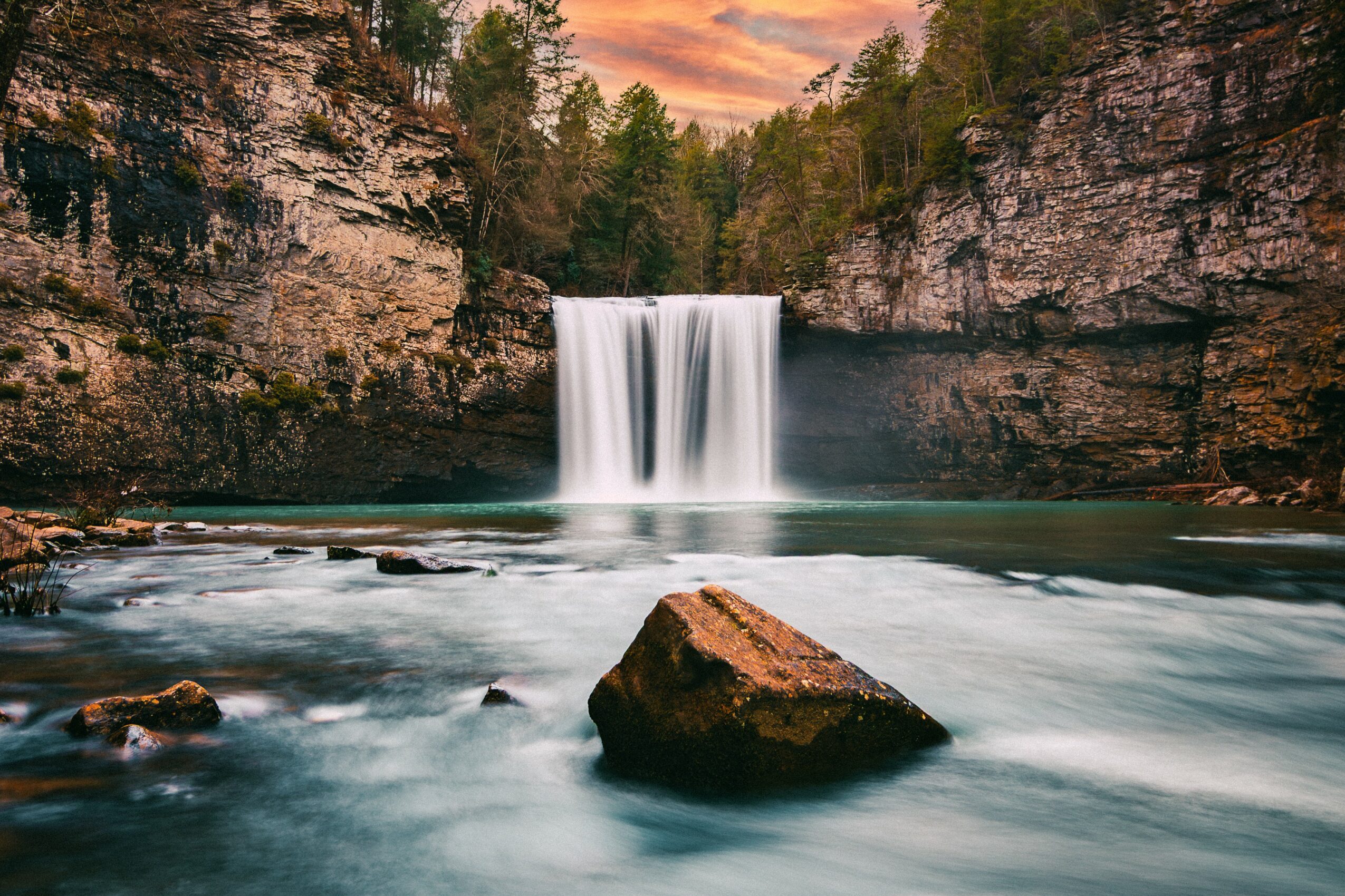 Waterfall flowing into a river between rocky cliffs with trees and a colorful sky in the background.