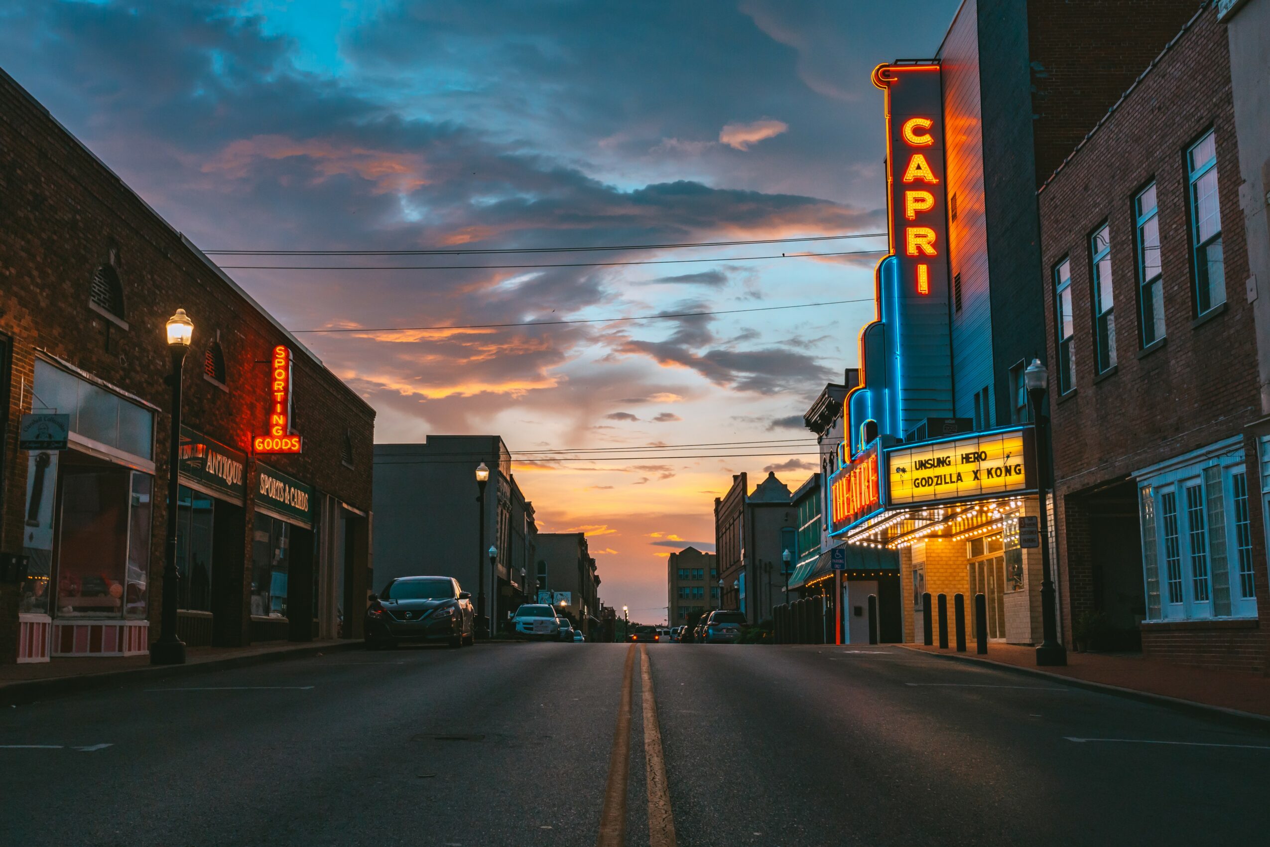 Street view at sunset with buildings, neon signs, and a cloudy sky, empty road, and distant horizon.