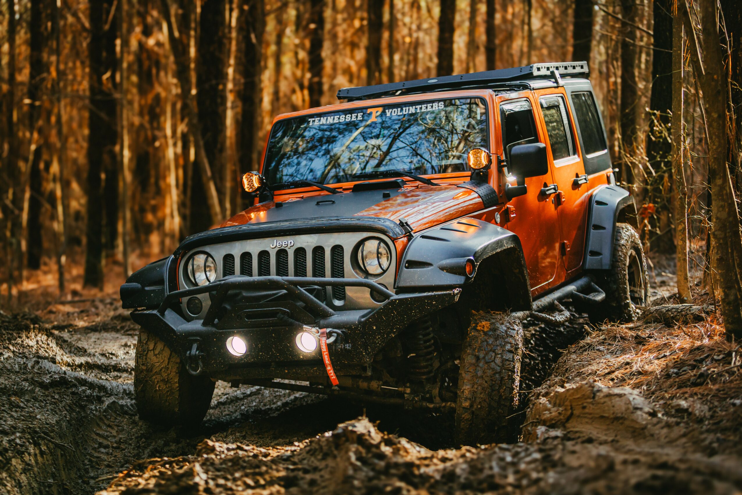 Orange and black off-road vehicle driving through a forest trail with trees and dirt, headlights on, mud on tires.