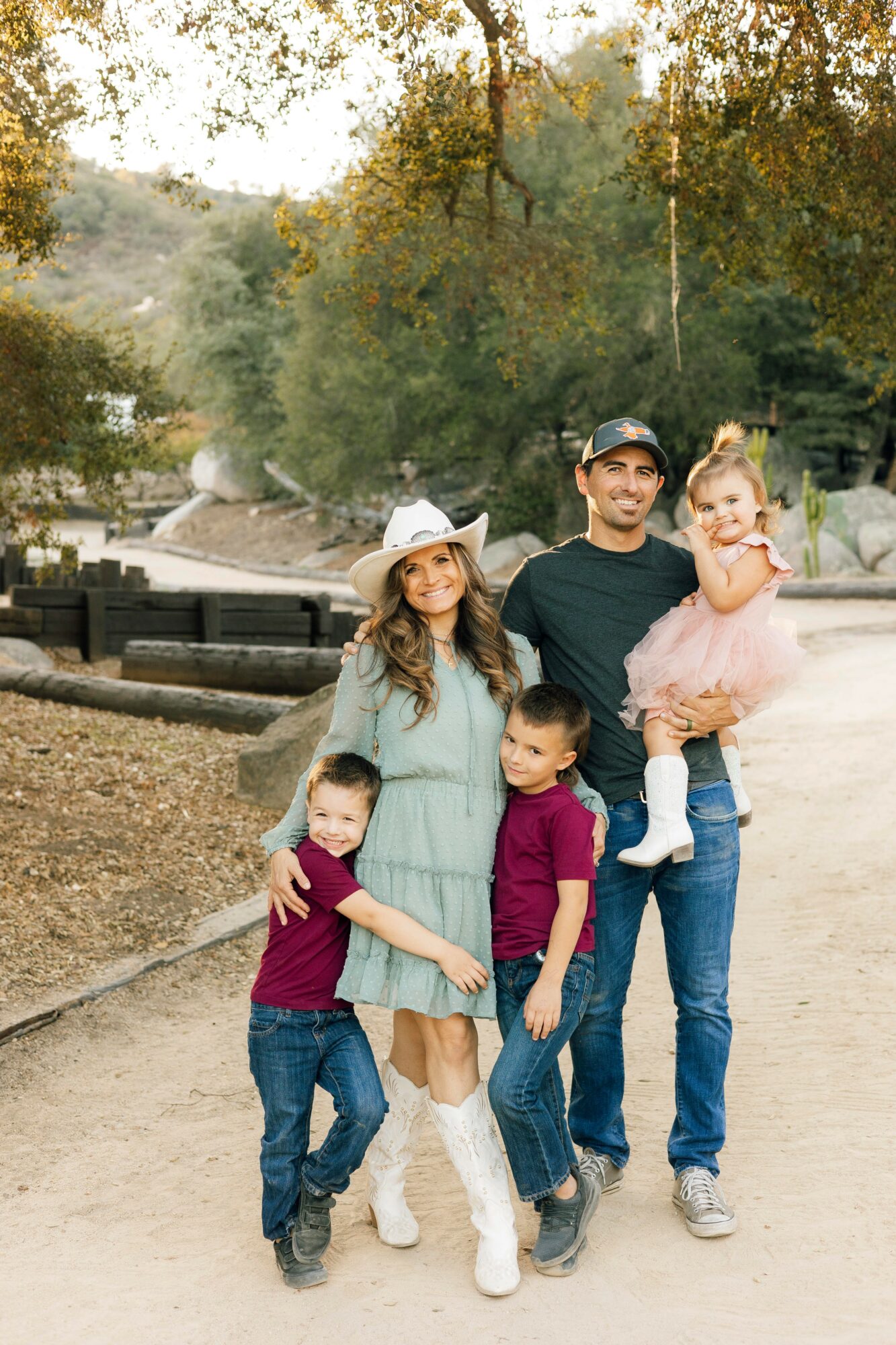 Family of five outdoors on a dirt path with trees, two children, a woman, and a man holding a young girl.