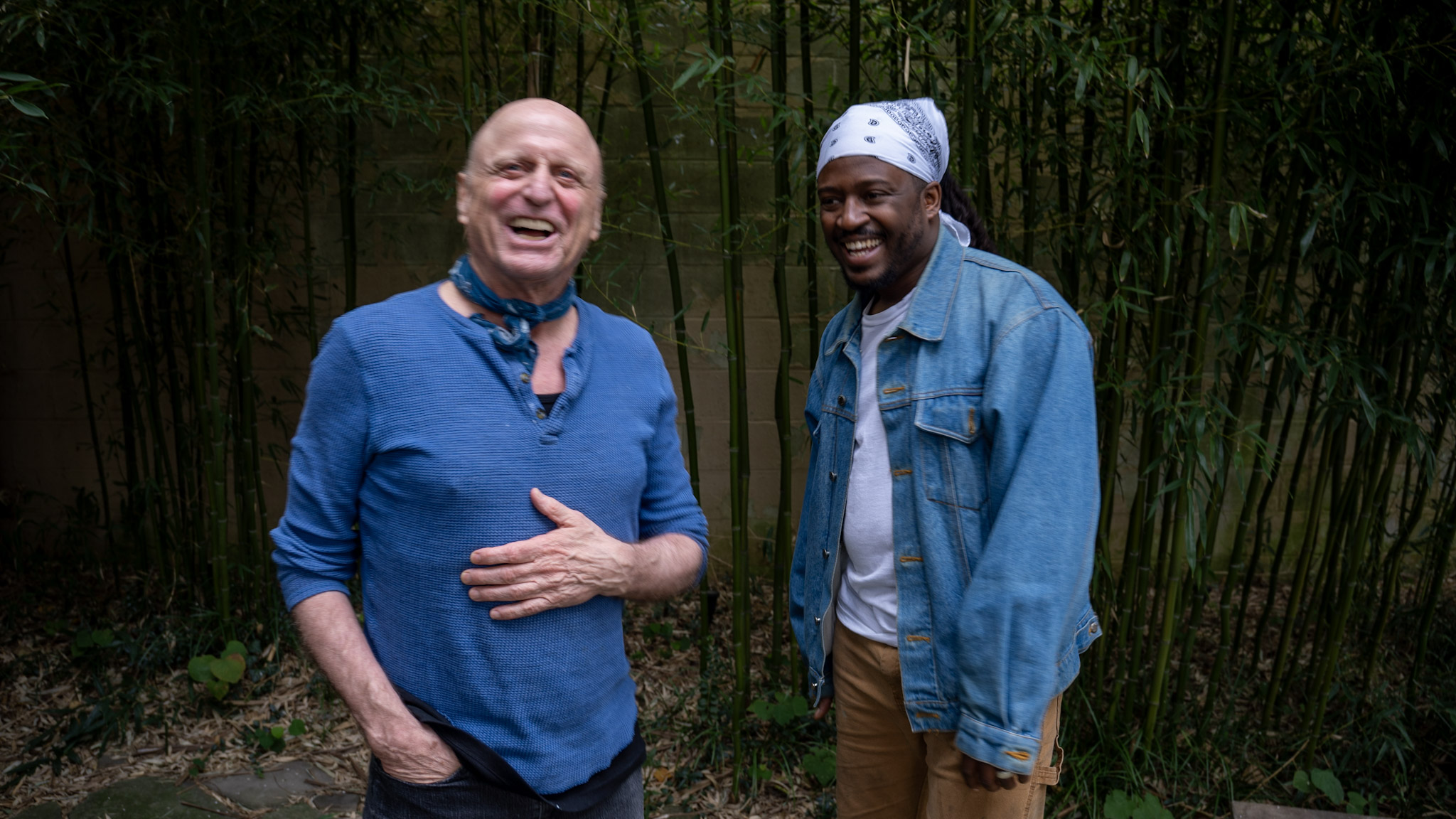 Two men smiling and laughing outdoors in front of bamboo plants, one wearing a blue shirt and the other a denim jacket.