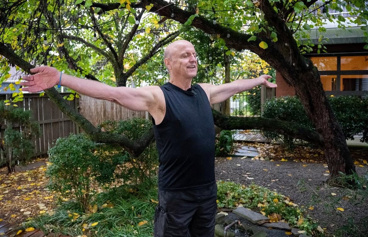 Man with arms outstretched standing outdoors in a garden with trees and bushes.