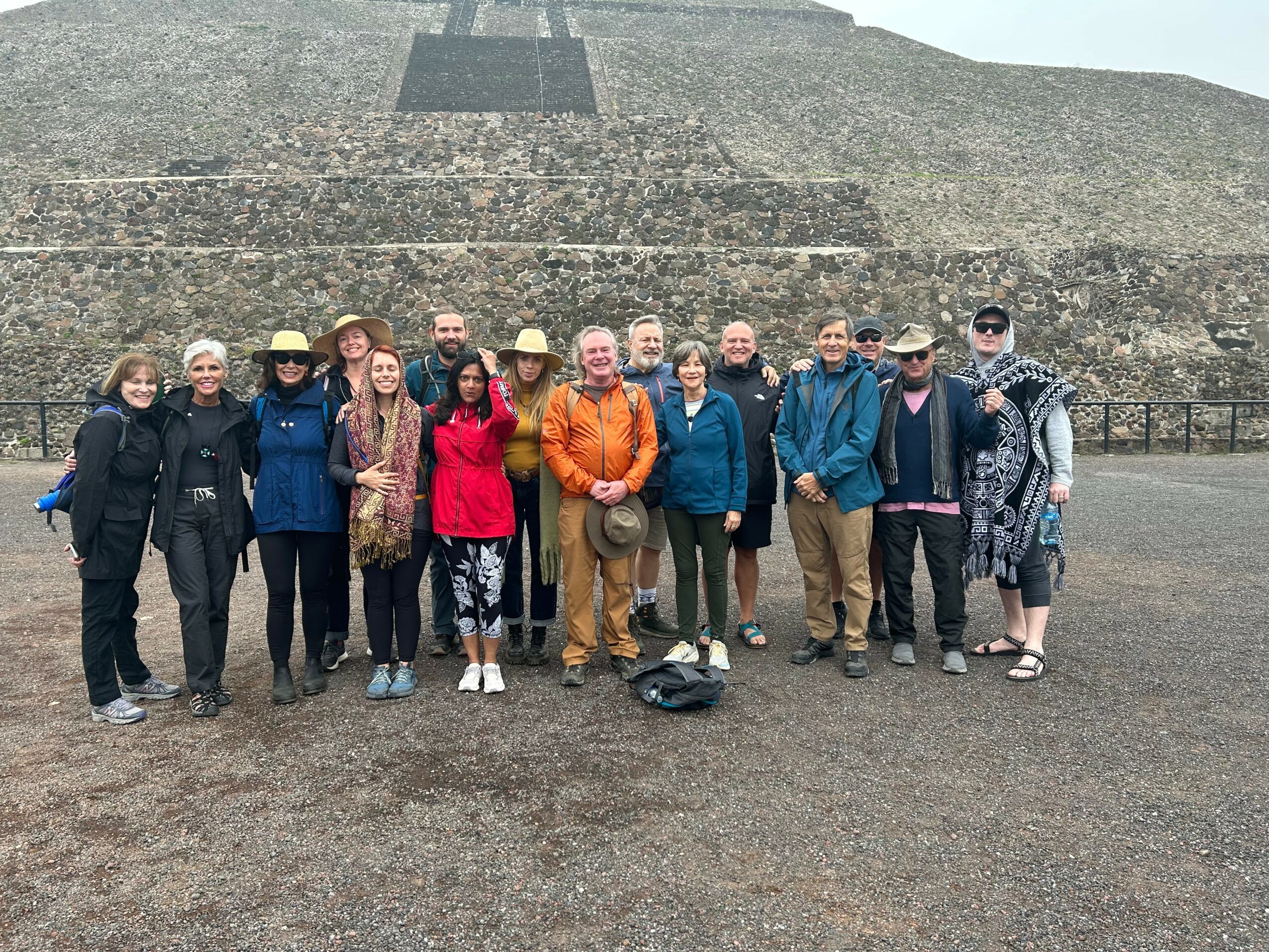 Group of 13 people standing in front of a stepped pyramid structure outdoors, some wearing hats and casual clothing.