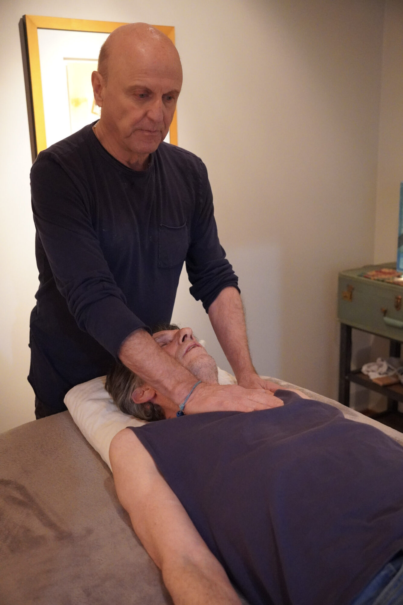 Man giving a massage to a person lying face down on a massage table in a room with artwork and furniture.