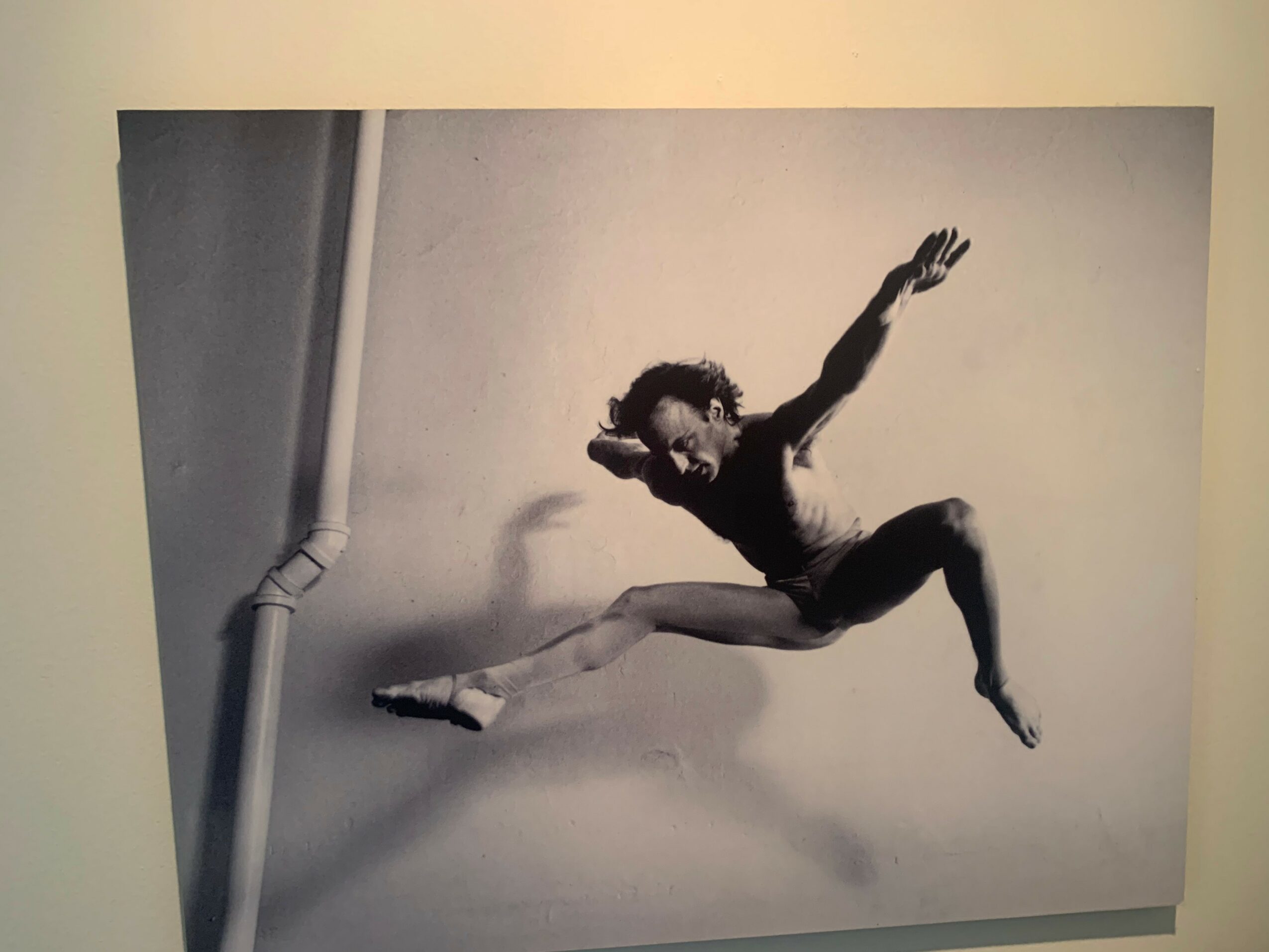 A person in mid-air jump with arms and legs extended, against a plain background with a pipe on the wall.
