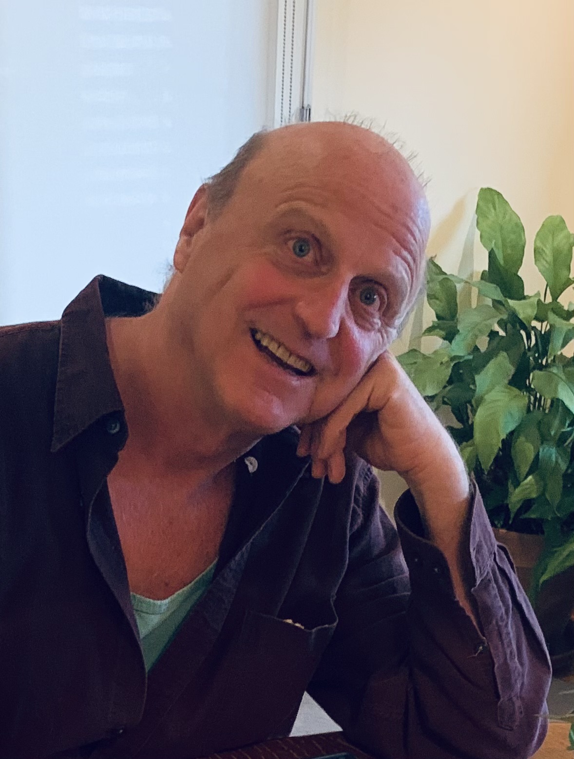 Man with a bald head smiling, resting his head on his hand, with green plants in the background.