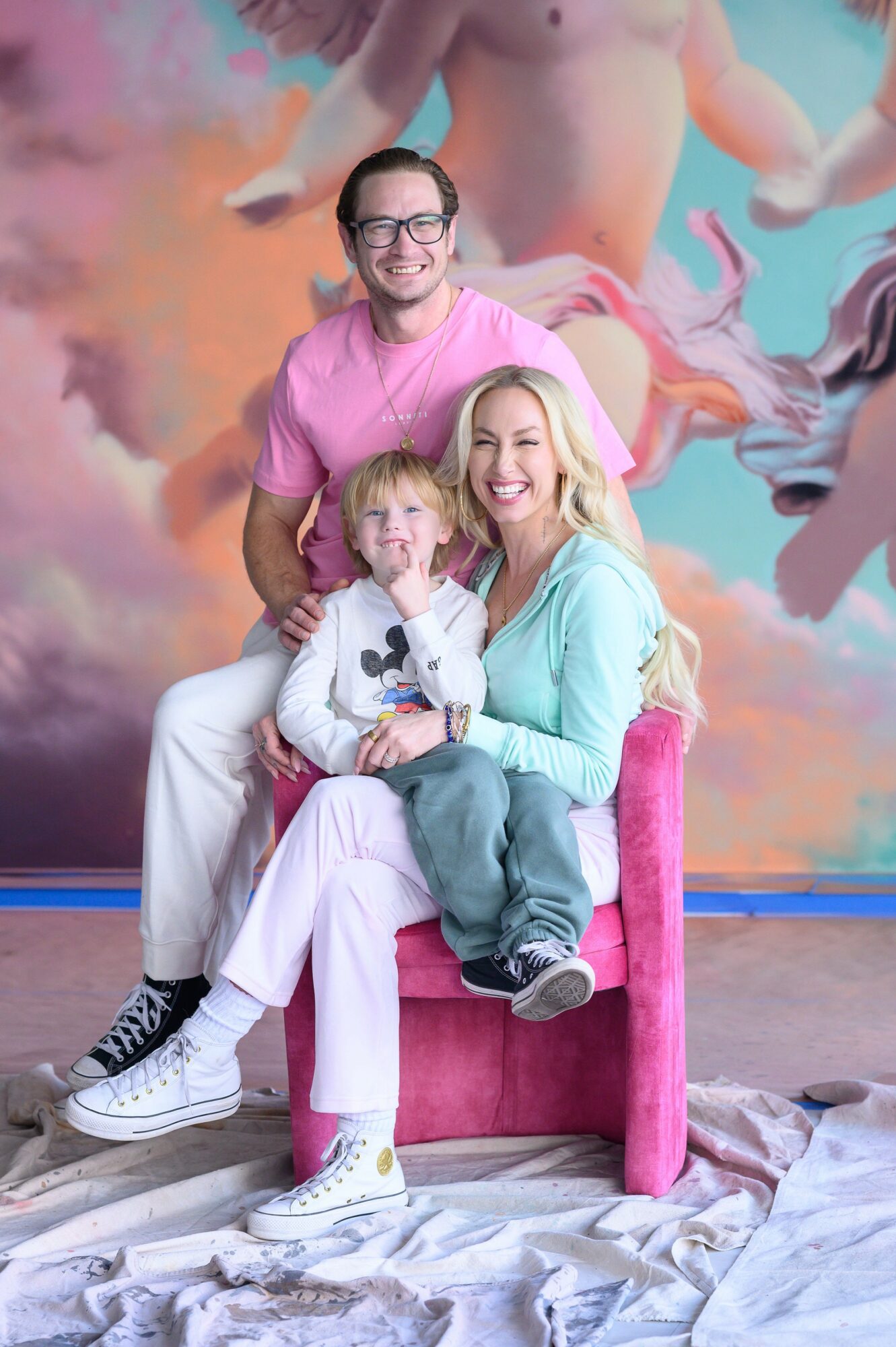 Three people, two adults and a child, sitting on a pink chair with a colorful mural background, smiling at the camera.