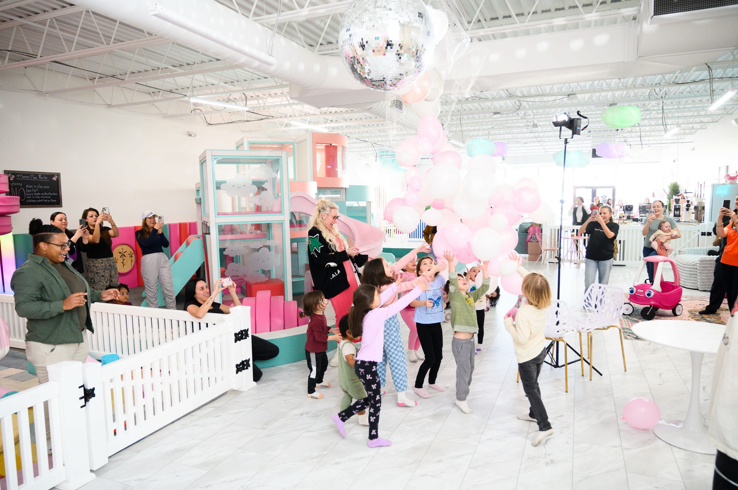 Children and adults enjoy a party with balloons, a disco ball, and decorations in a bright indoor space.