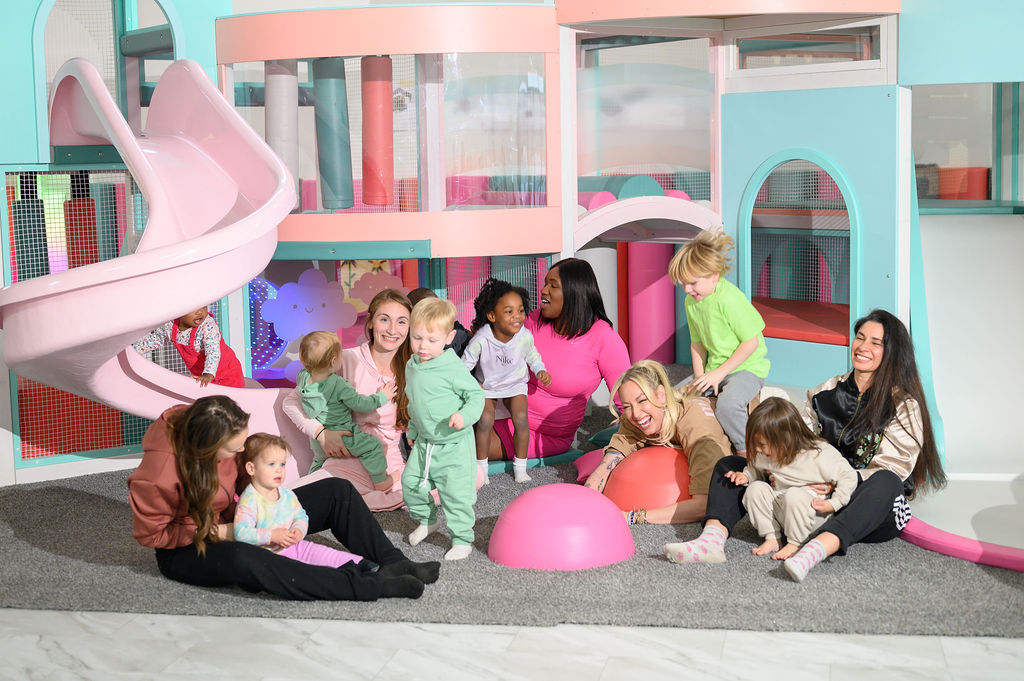 Group of children and adults playing on a soft mat in a colorful indoor playground, with slides and play structures in the background.