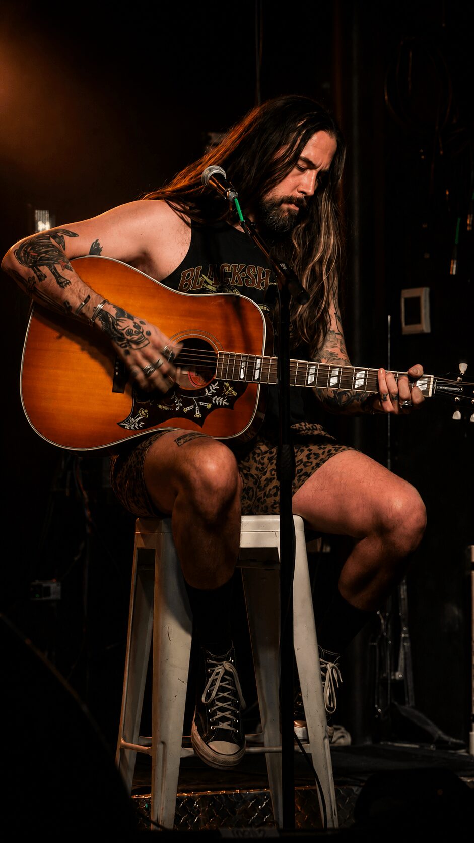 Person with long hair and tattoos playing an acoustic guitar on stage, seated on a stool, in a dark setting.