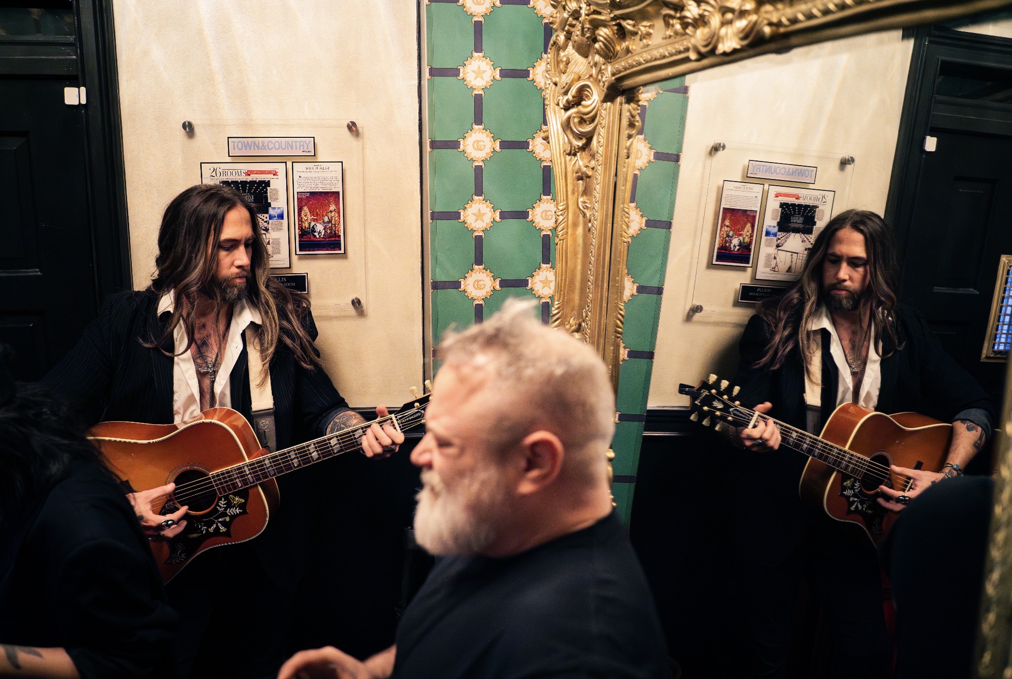 Two women playing guitars in a decorated room with a large mirror and ornate gold frame, and a man with a beard in foreground.