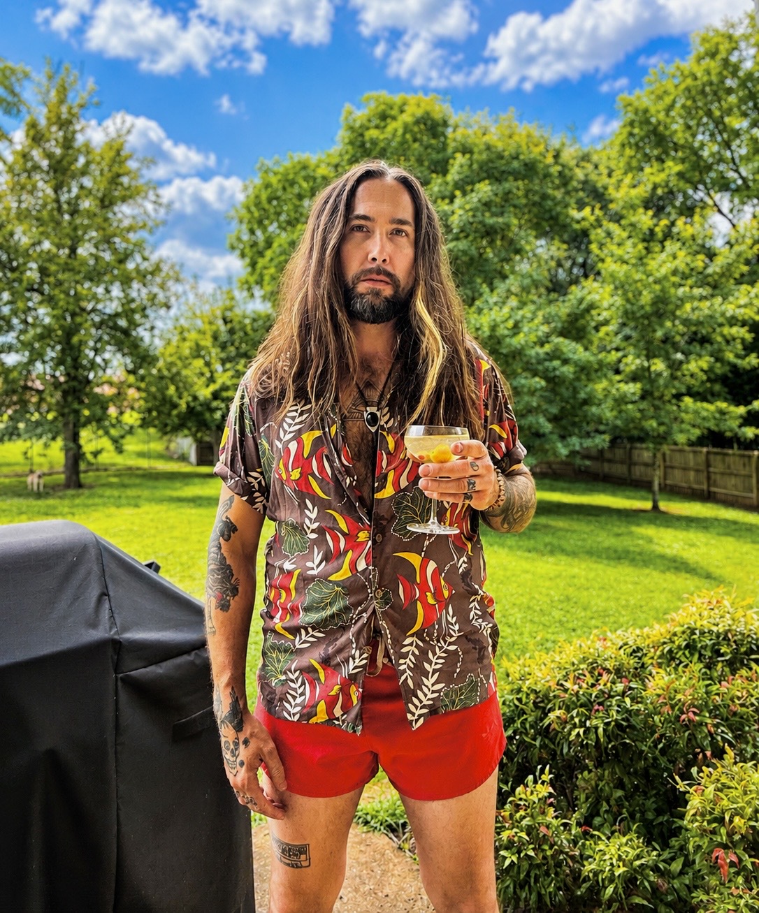 Man with long hair and beard holding a drink outdoors in a park with green trees and blue sky