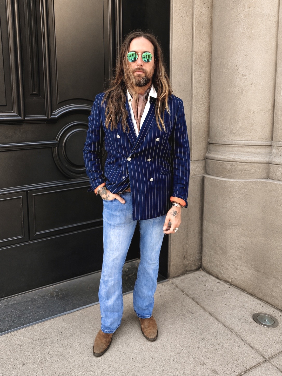 Man with long hair and sunglasses standing outside near a black door and stone wall, wearing a blue pinstripe blazer and jeans.