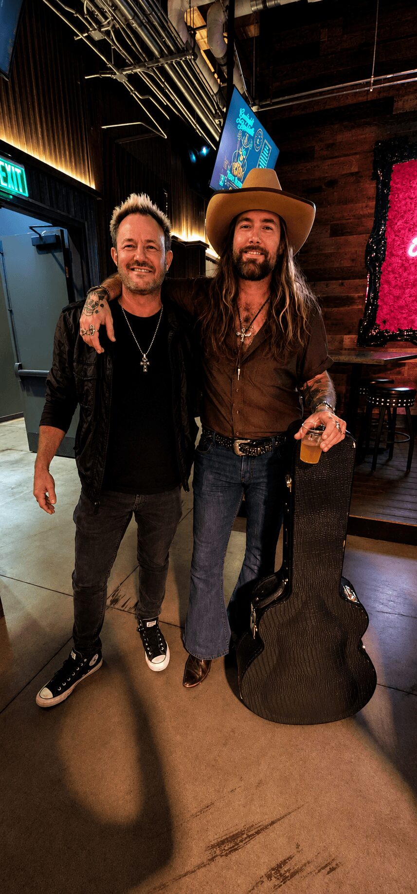 Two men standing together indoors, one with a guitar case, smiling, with a dark background and colorful decorations.