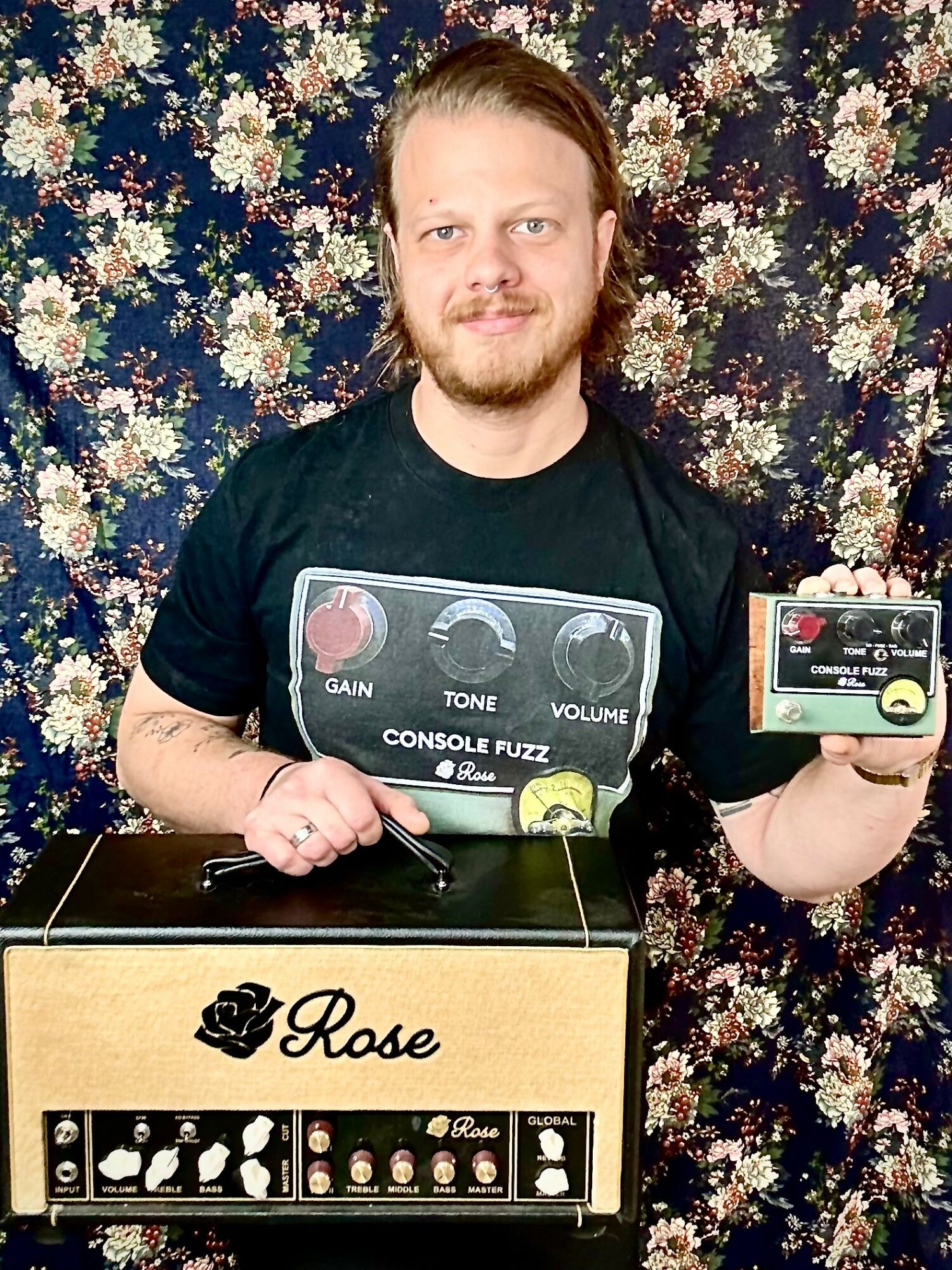Person with long hair and beard holding a small box, standing behind a guitar effects pedal and a box labeled 'Rose', against a floral background.