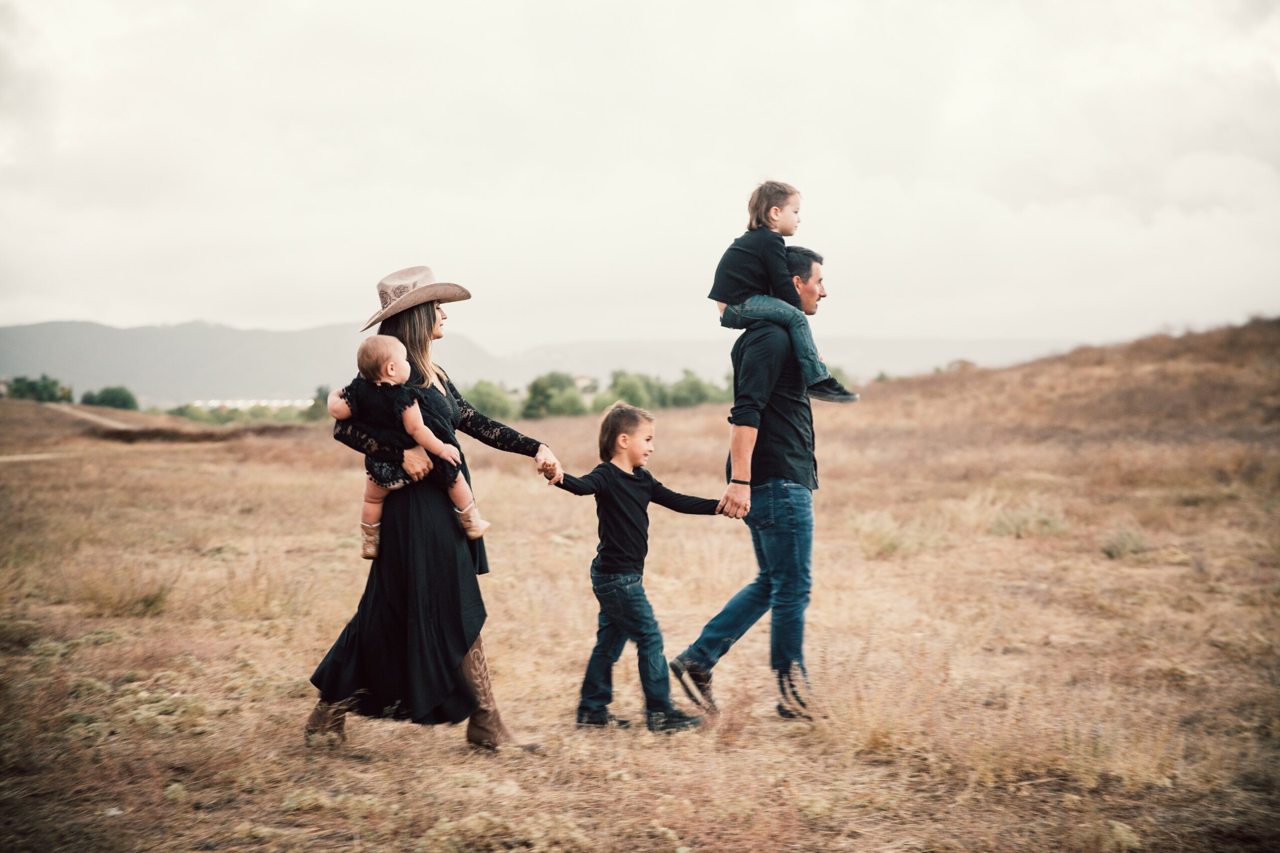 Family of five walking across a dry field, with a woman in a hat, children, and a man carrying a child on his shoulders.