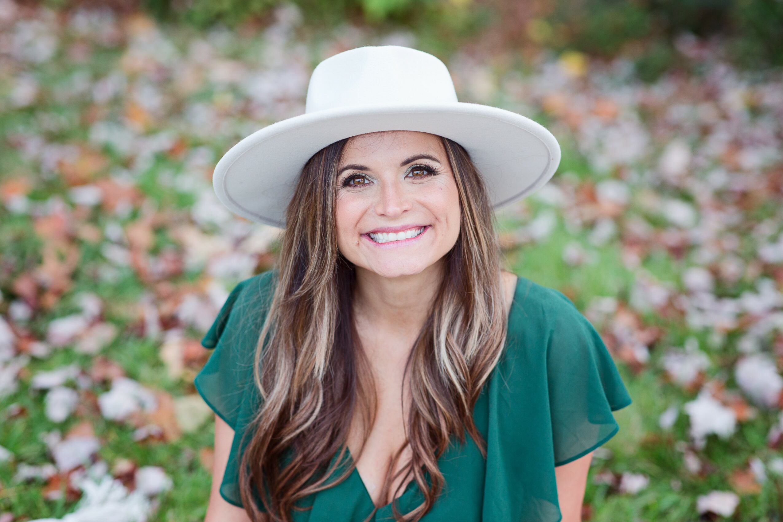 Young woman with long hair wearing a white hat and green top, smiling outdoors with colorful flowers in background.
