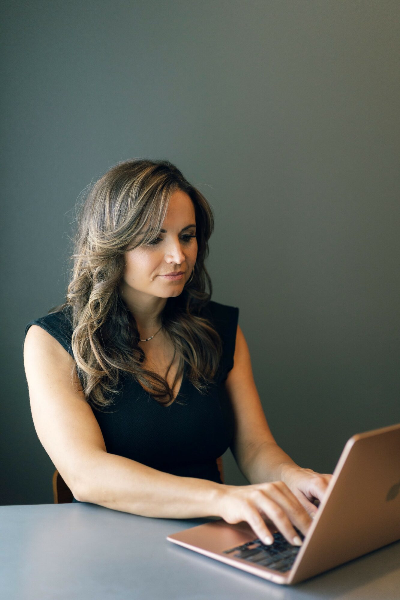 Woman with wavy hair working on a laptop at a desk against a plain wall.
