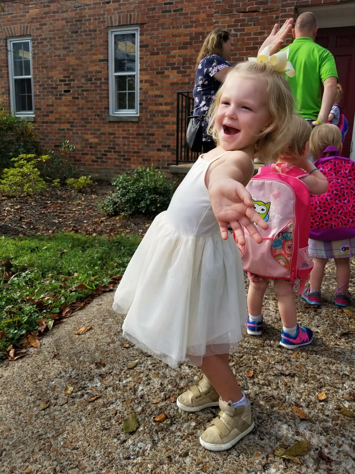 Young girl in a white dress making a peace sign, standing outdoors on a gravel path with children in the background.