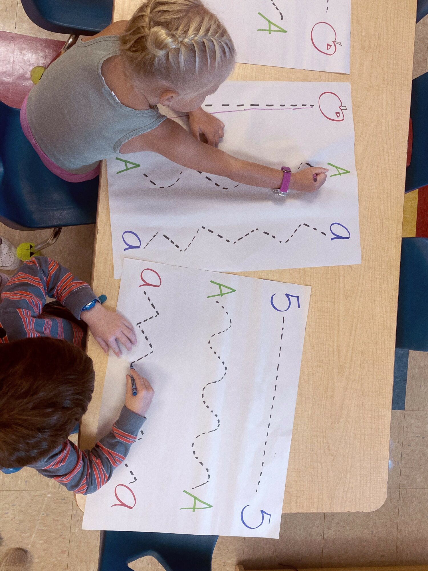 Two children draw zigzag lines on large paper with colorful markers, practicing writing patterns.
