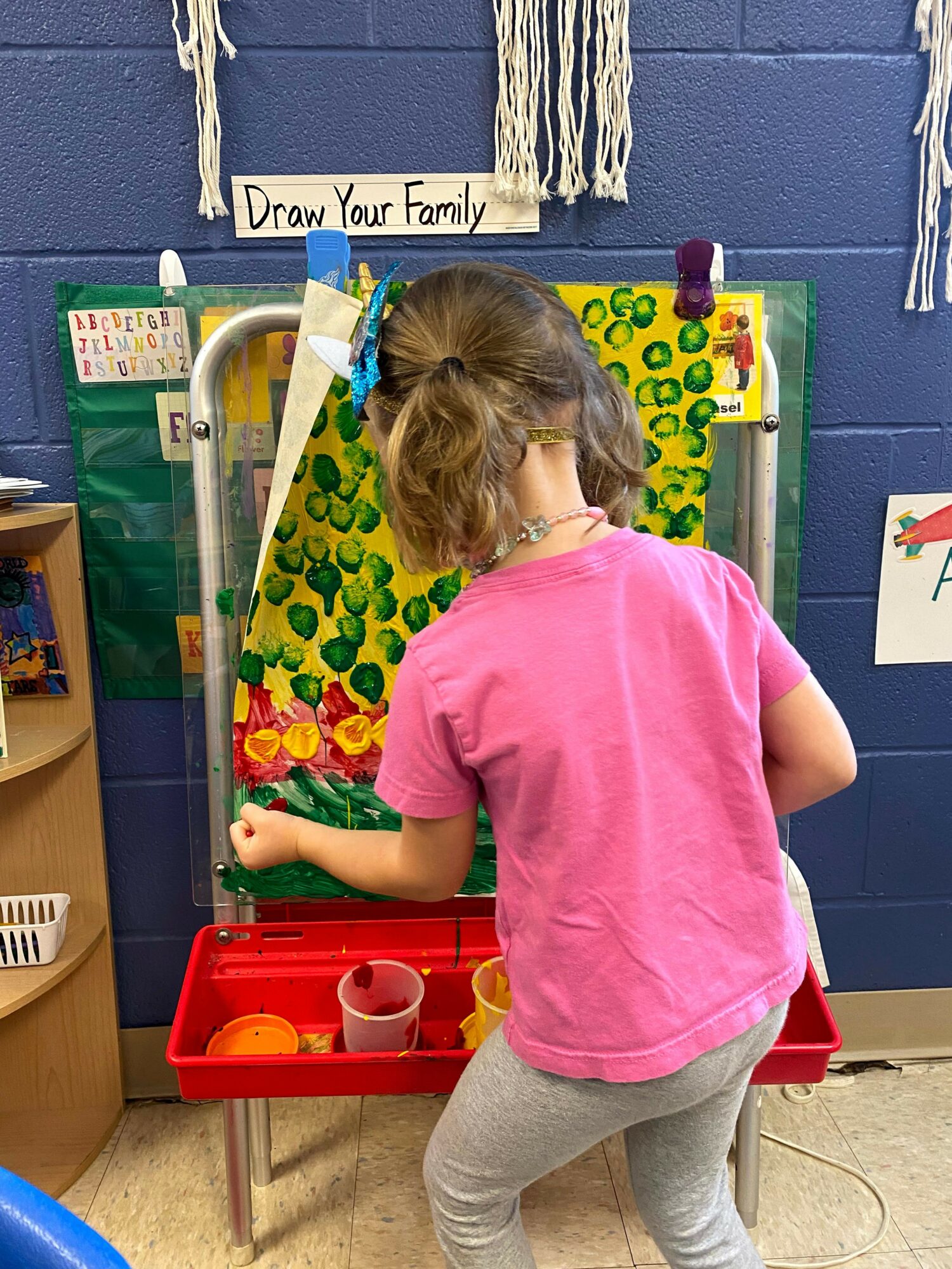 Child in pink shirt and gray pants playing with a toy kitchen set in a classroom.