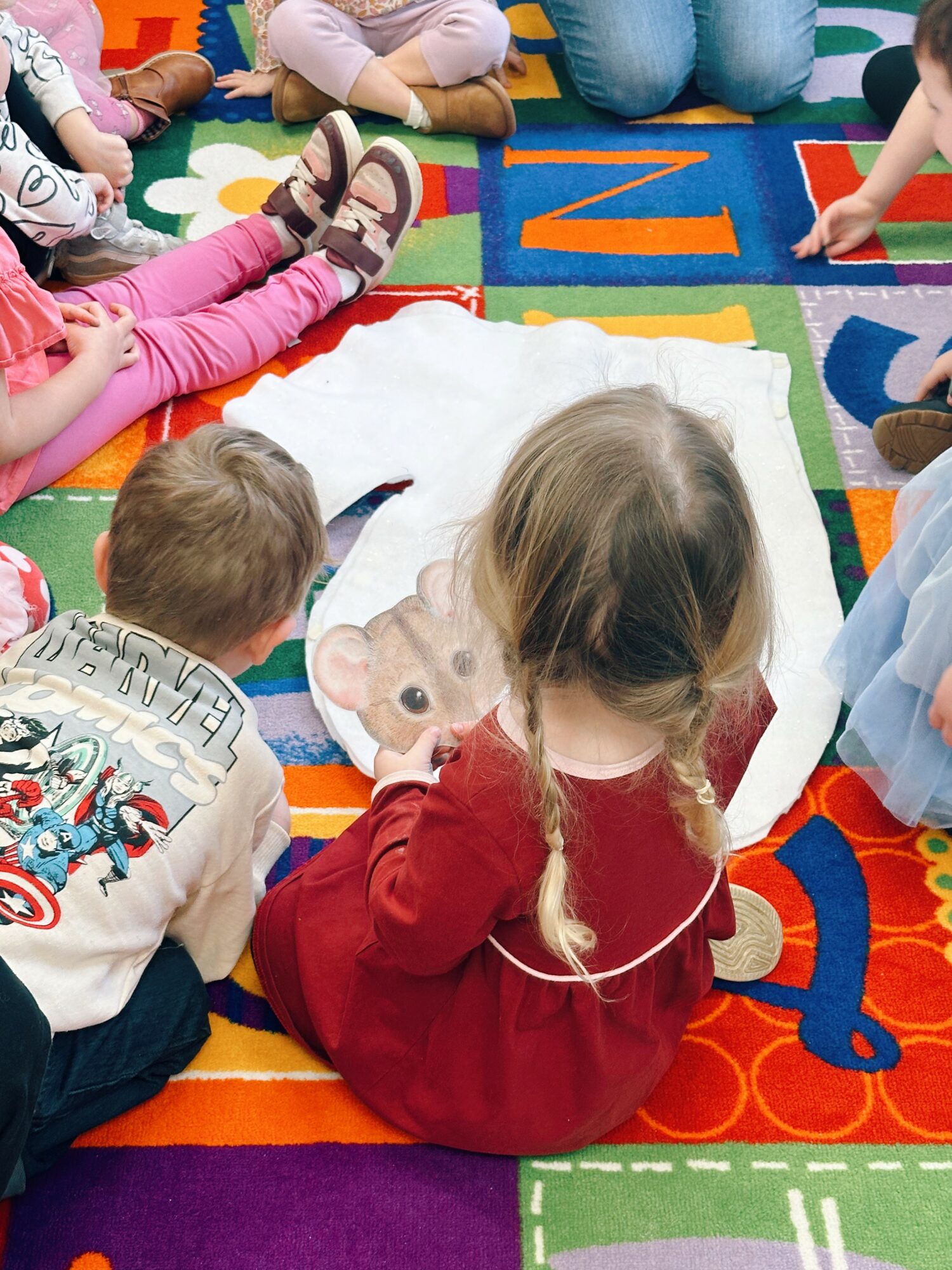 Children sitting on a colorful alphabet rug, gathered around a white cloth with a bear face, engaging in a group activity.