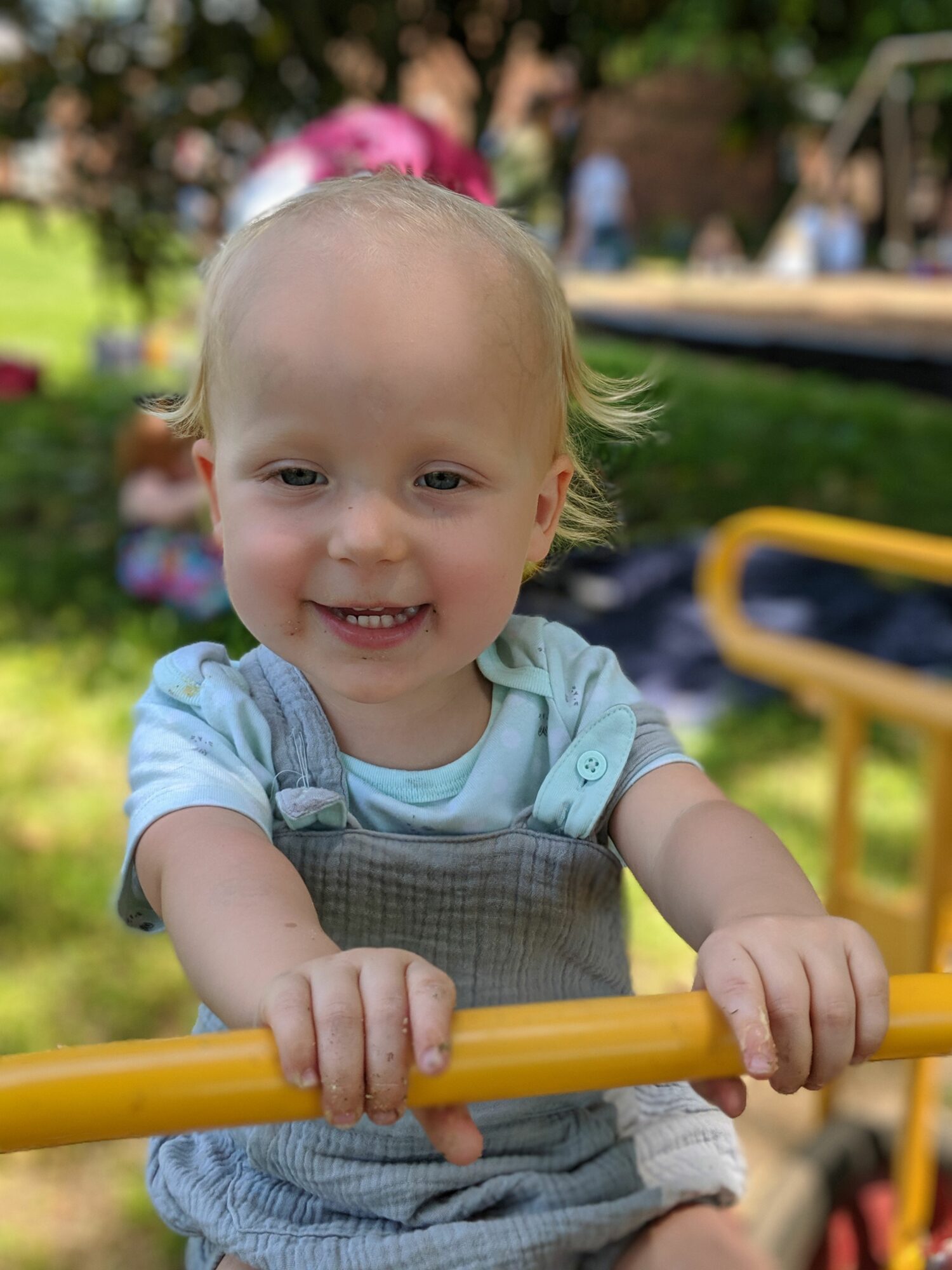 Young child with blonde hair smiling, holding yellow playground equipment, outdoors with blurred background of people and trees.