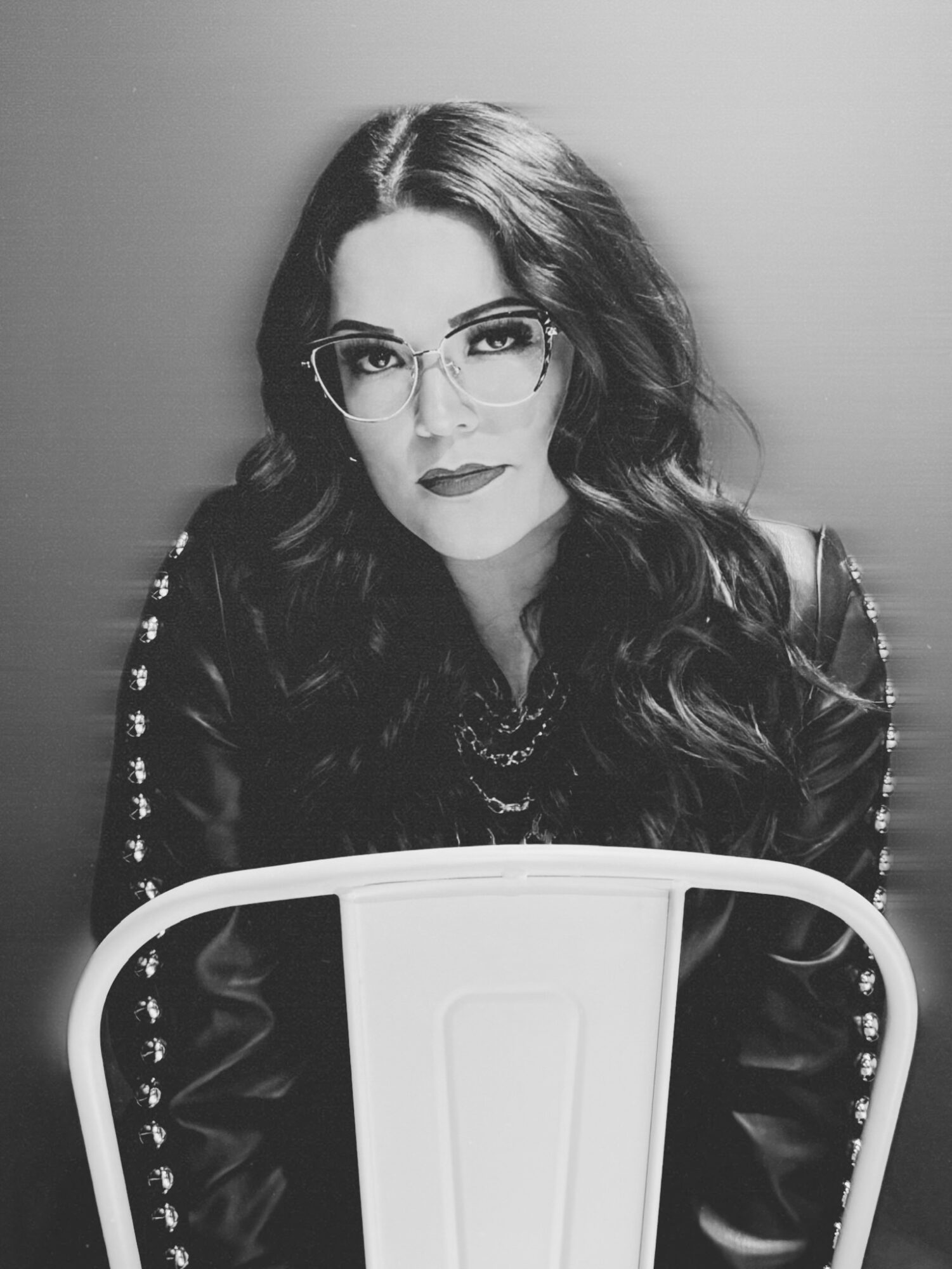 Woman with wavy hair, glasses, and leather jacket, sitting behind a chair, looking at the camera, in black and white.