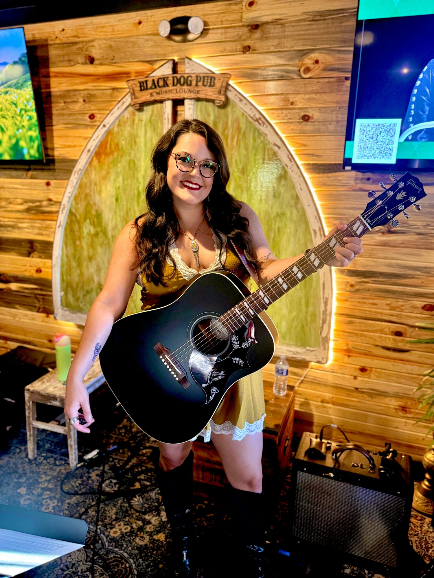 Woman with glasses holding a black acoustic guitar, smiling, standing in front of a wooden wall with a sign that says 'Black Dog Pub'.
