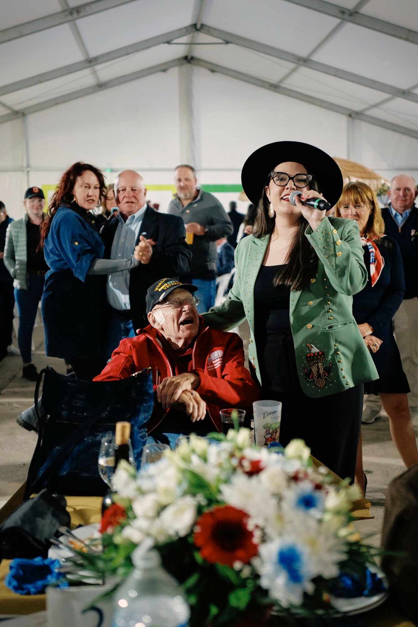 Group of people gathered indoors, woman with glasses and hat smiling, table with flowers in foreground.