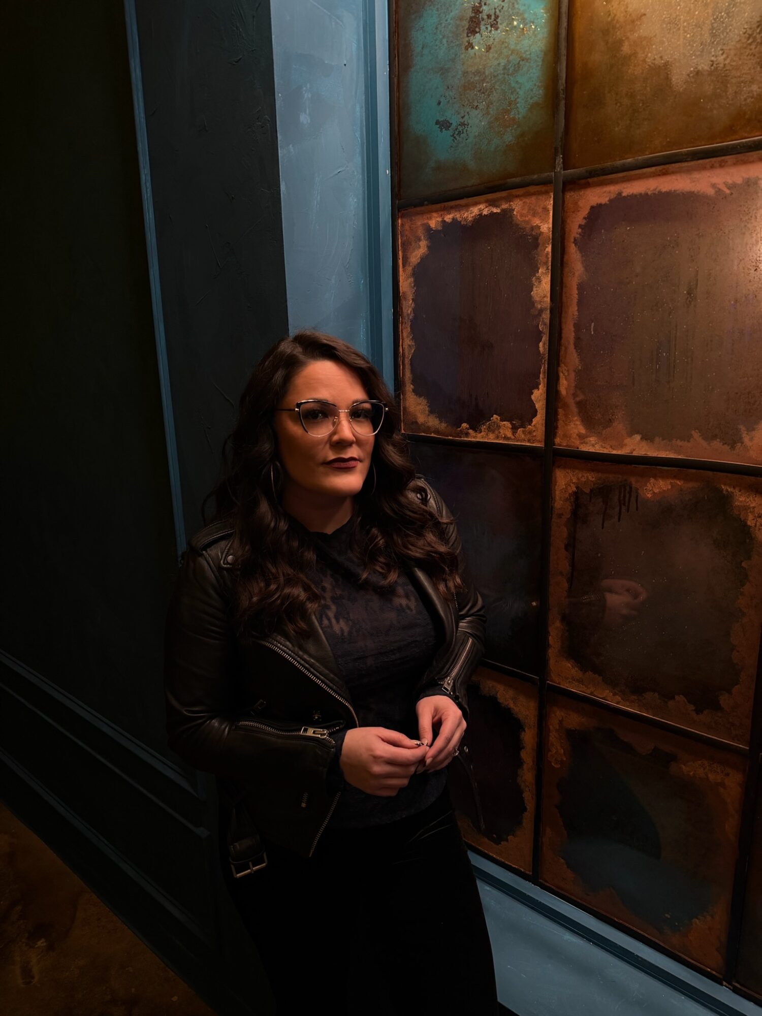 Woman with long dark hair and glasses standing in front of a rusted metal wall, wearing a black leather jacket.