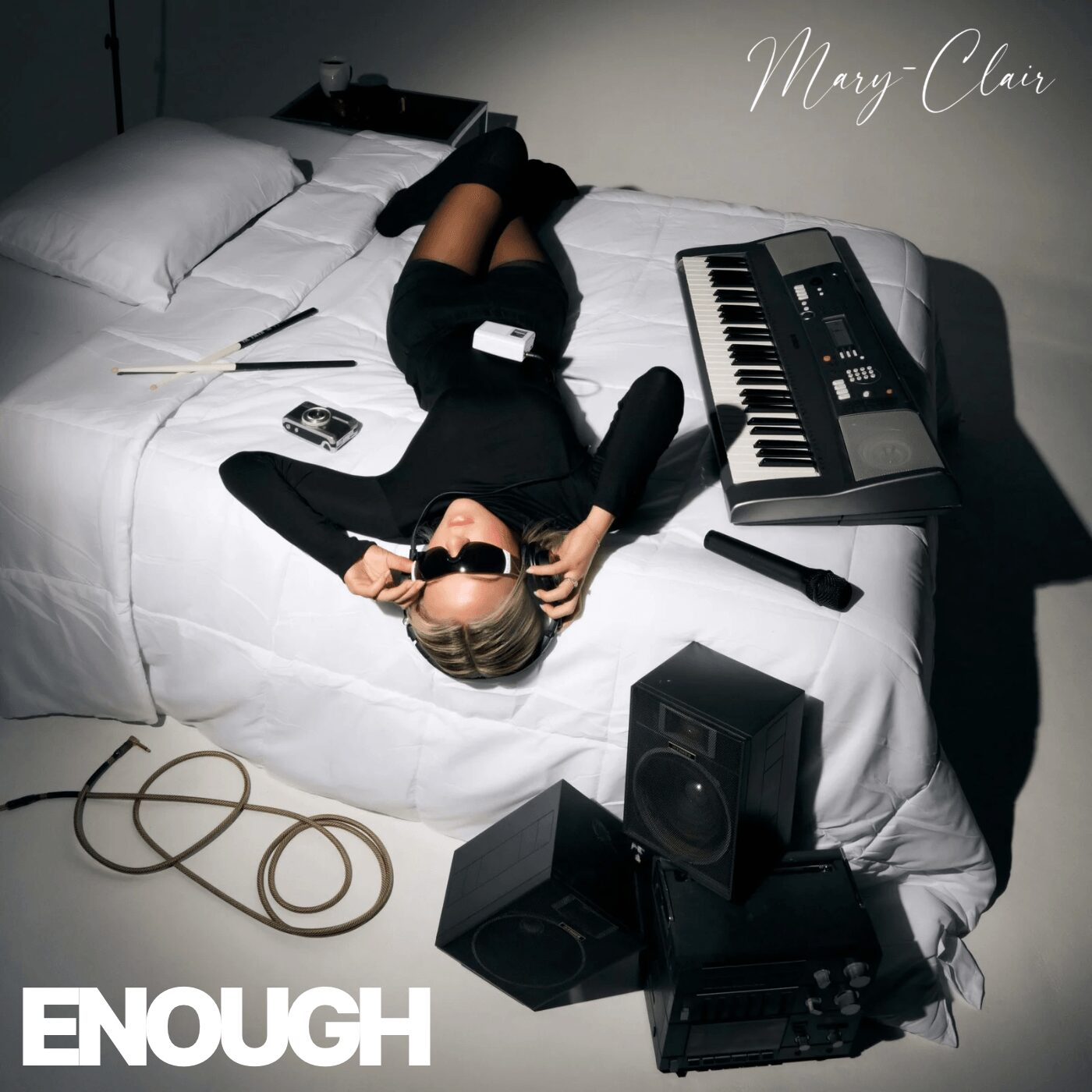 Person lying on bed with sunglasses, surrounded by music equipment, in a room with white walls and bedding.
