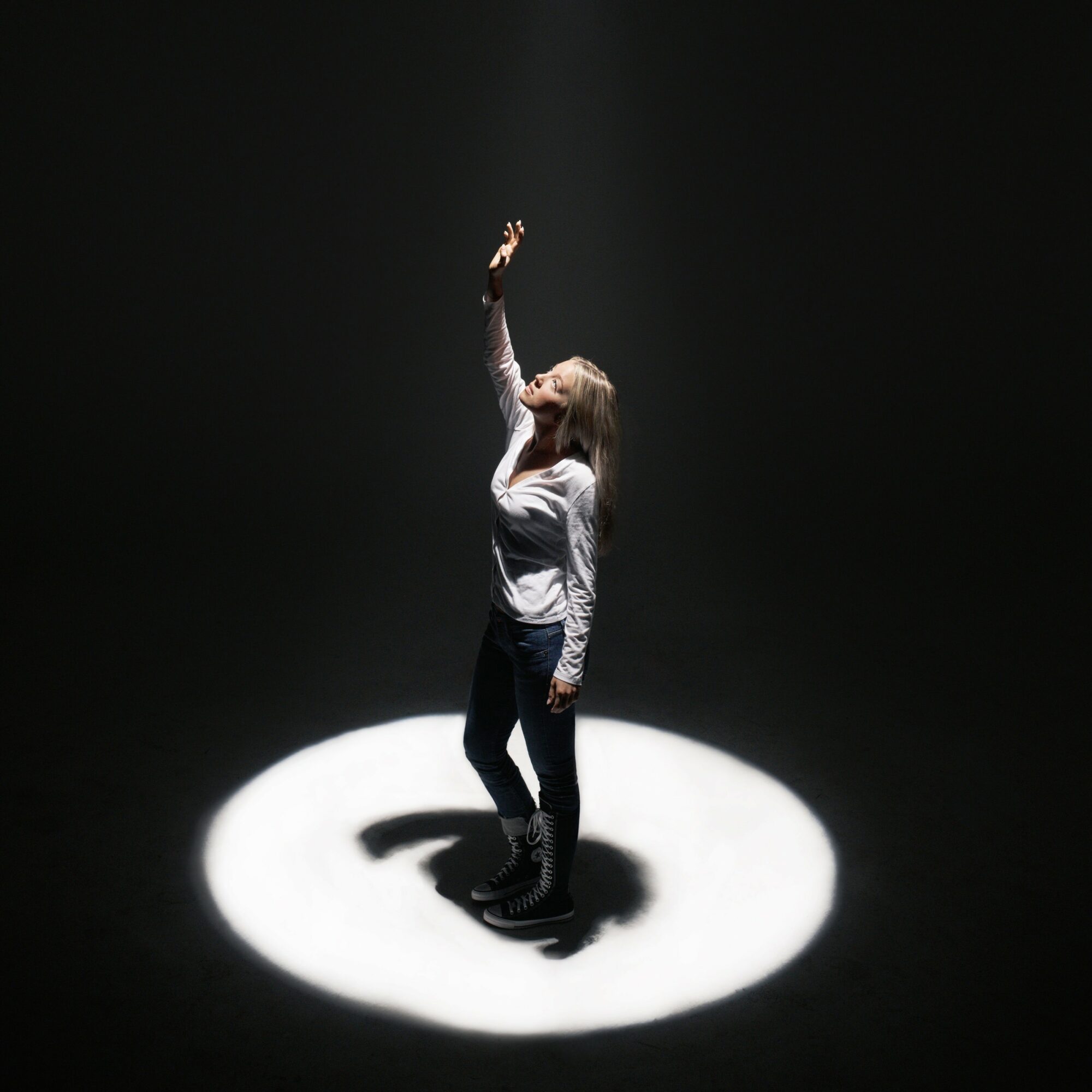 Person standing in a spotlight, reaching upward with one hand, surrounded by darkness.