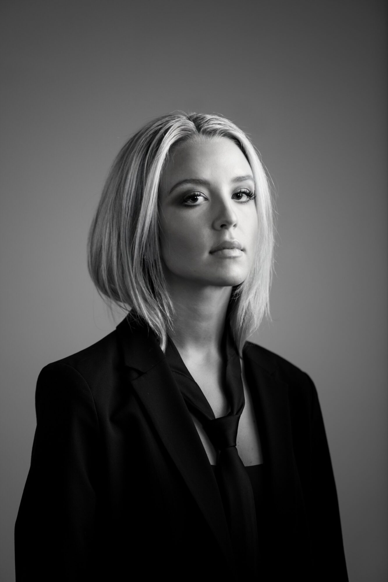 Portrait of a woman with shoulder-length hair, wearing a dark blazer, looking at the camera against a plain background.