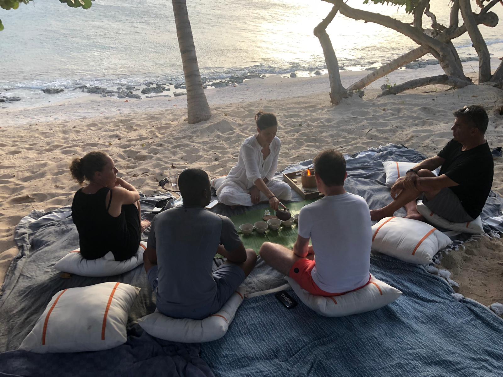 Group of five people sitting on cushions and a blanket on the beach, with trees and water in the background.