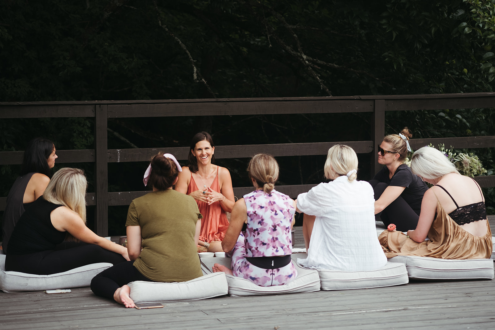 Group of eight women sitting on cushions outdoors, engaged in conversation, with a wooden fence and trees in background.