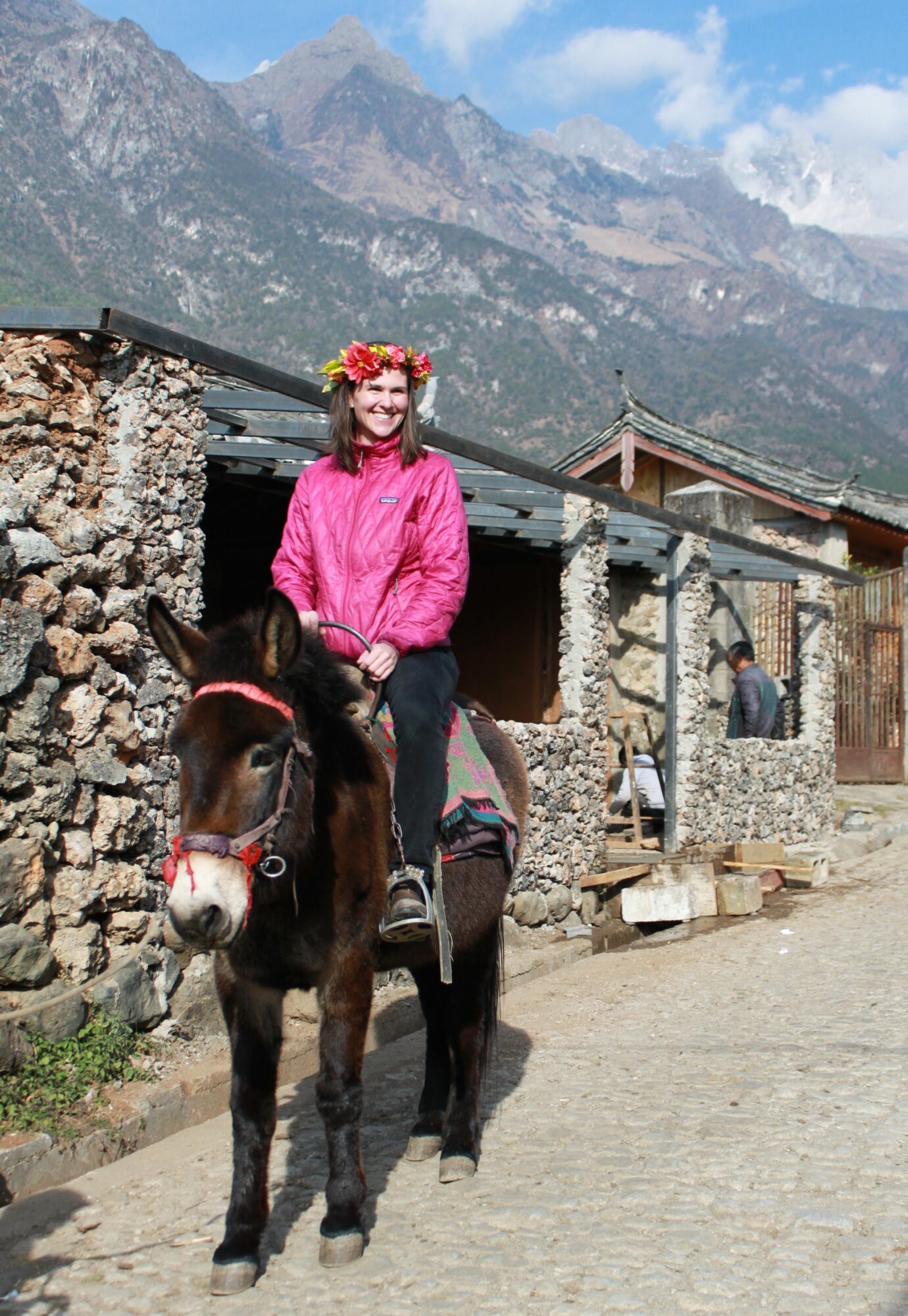 Woman wearing a pink jacket and flower crown riding a small horse on a cobblestone path with stone buildings and mountains in background.