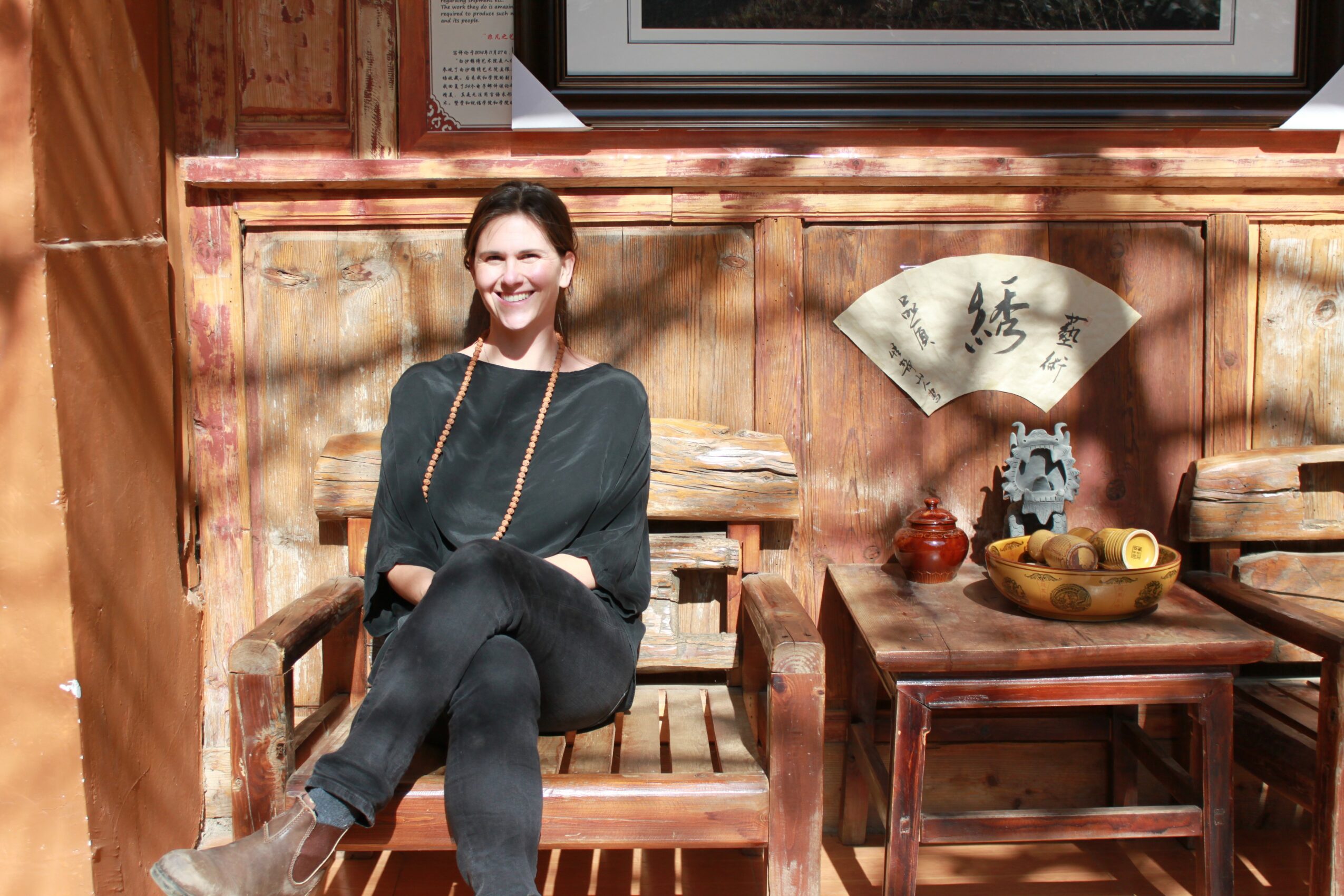 Woman sitting on a wooden bench outdoors, smiling, with a decorative fan and table behind her.