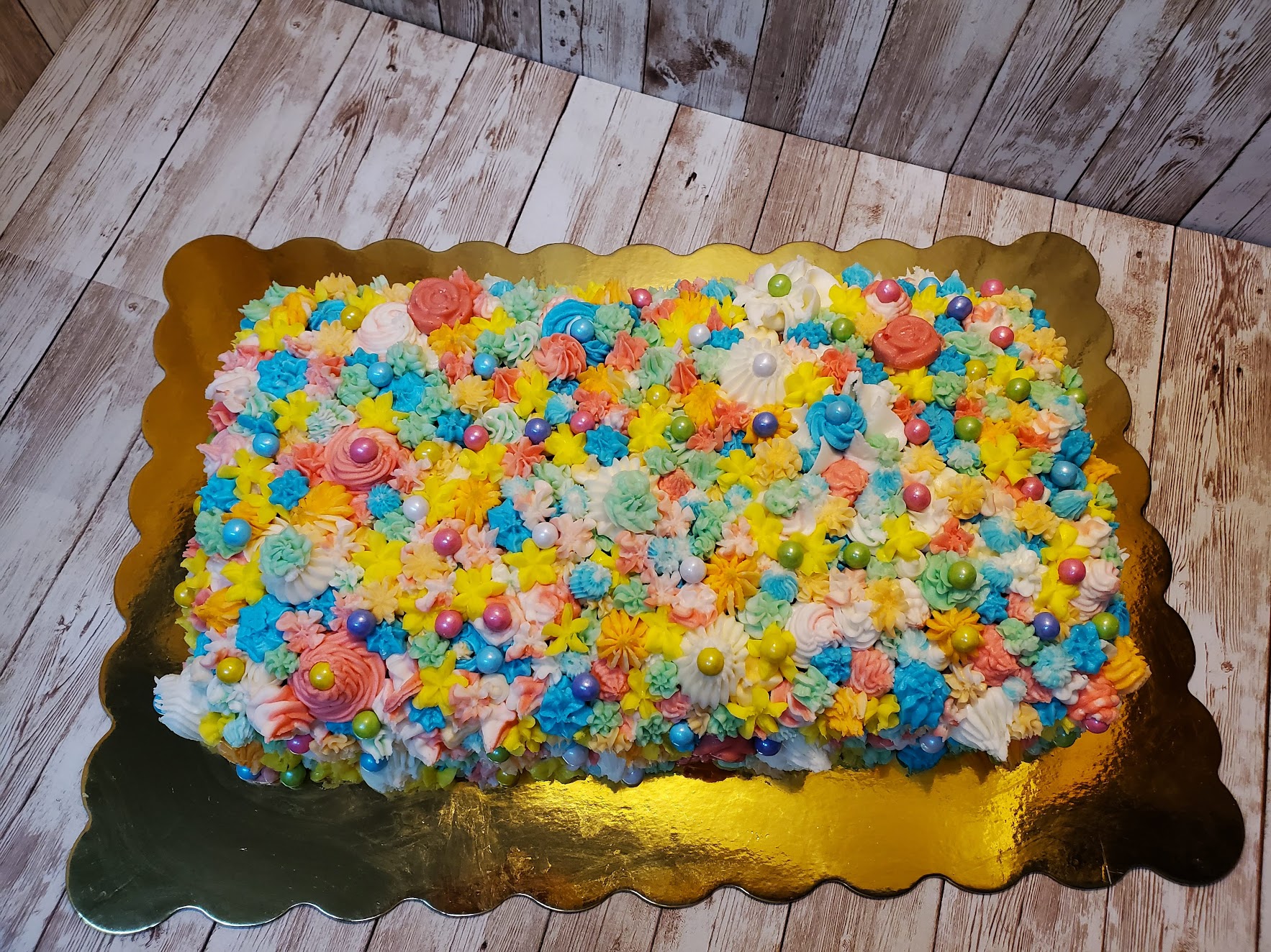 Colorful decorated rectangular cake with flowers and sprinkles on a gold tray, on a wooden surface.