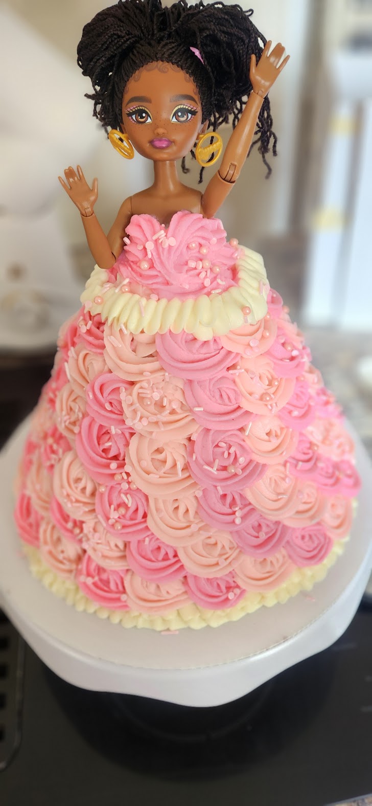Decorative doll with dark hair in a pink and cream dress, standing on a cake with pink and cream frosting rosettes.