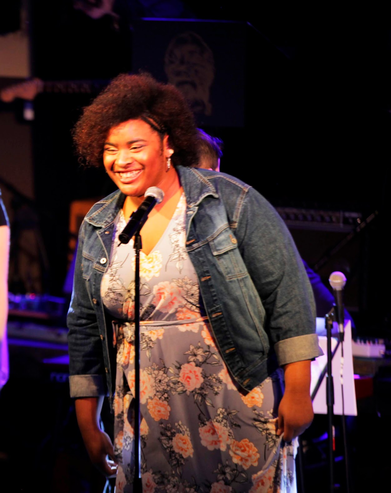 Smiling woman with curly hair in a floral dress and denim jacket speaking into a microphone on stage.