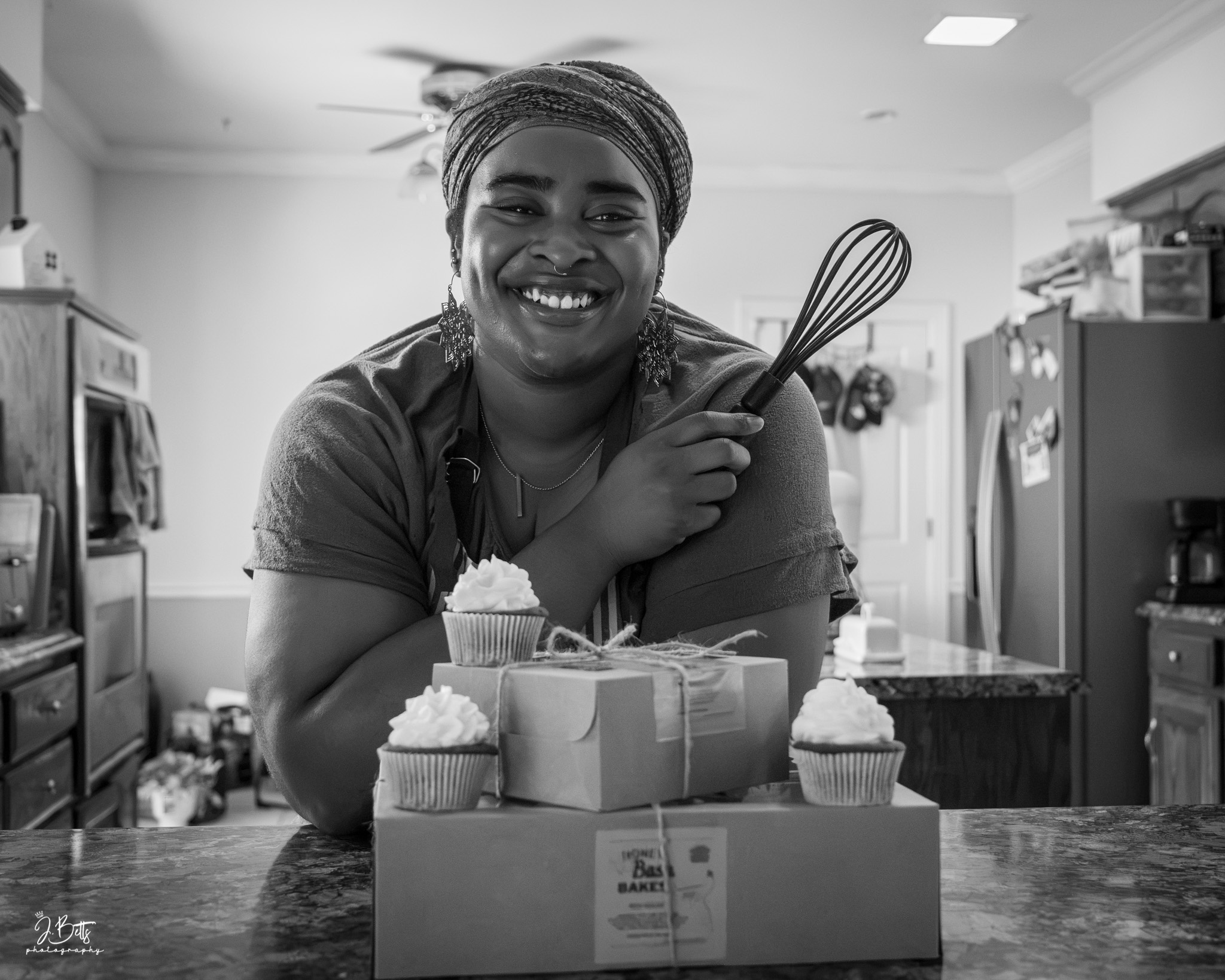 Smiling woman holding a whisk in a kitchen, with cupcakes and a gift box on the counter.