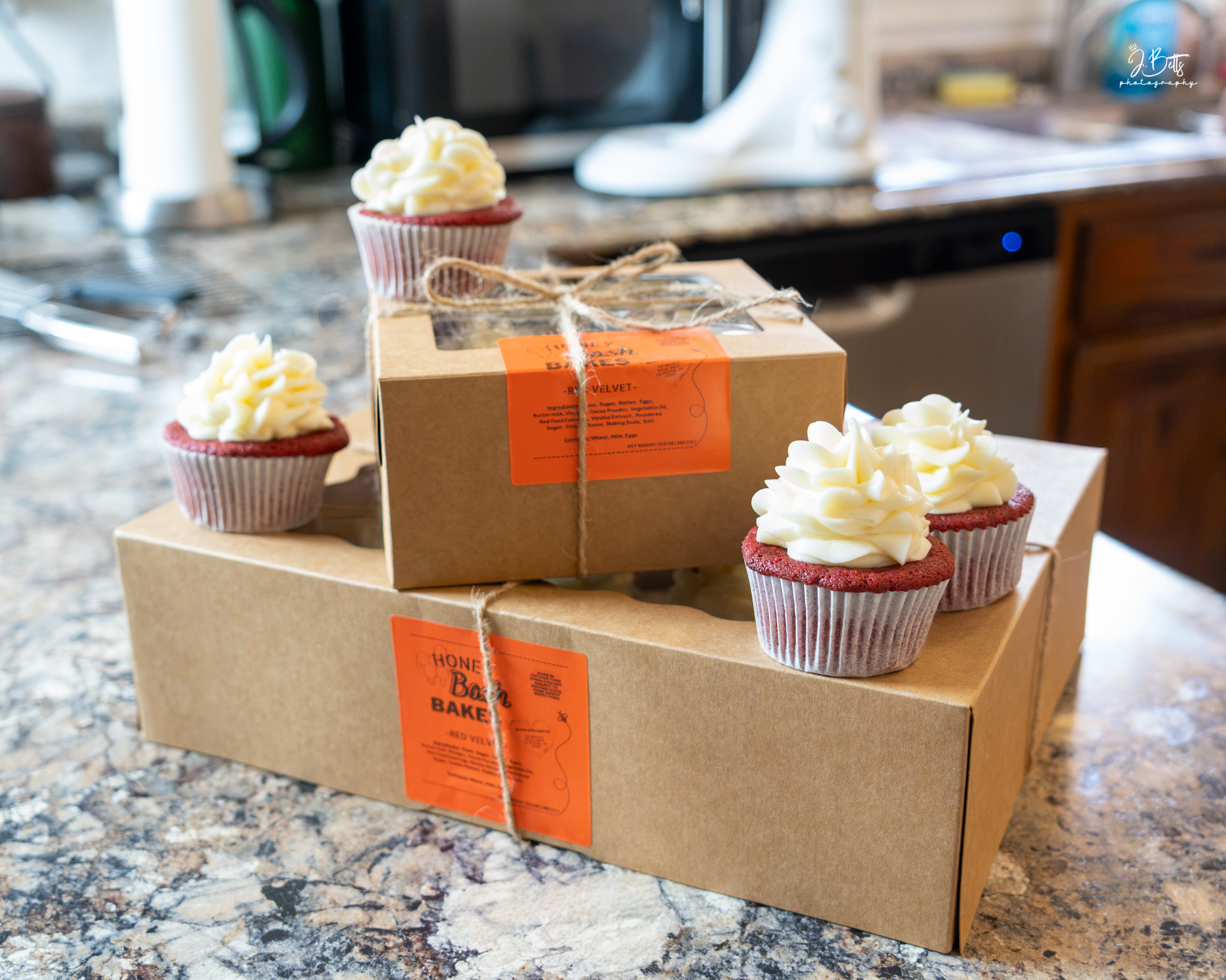 Three cupcakes with white frosting on two boxes on a kitchen countertop, with a microwave and kitchen items in the background.