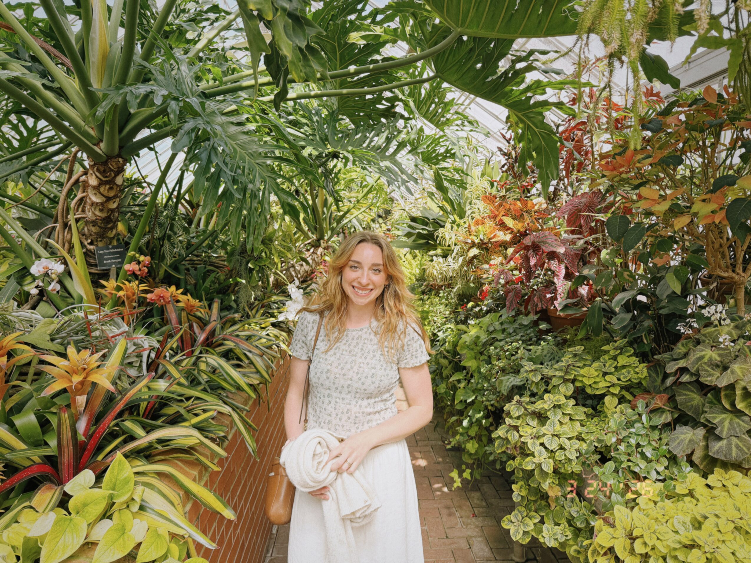 Young woman with curly hair smiling in a lush garden, holding a white hat and bag, surrounded by green plants.